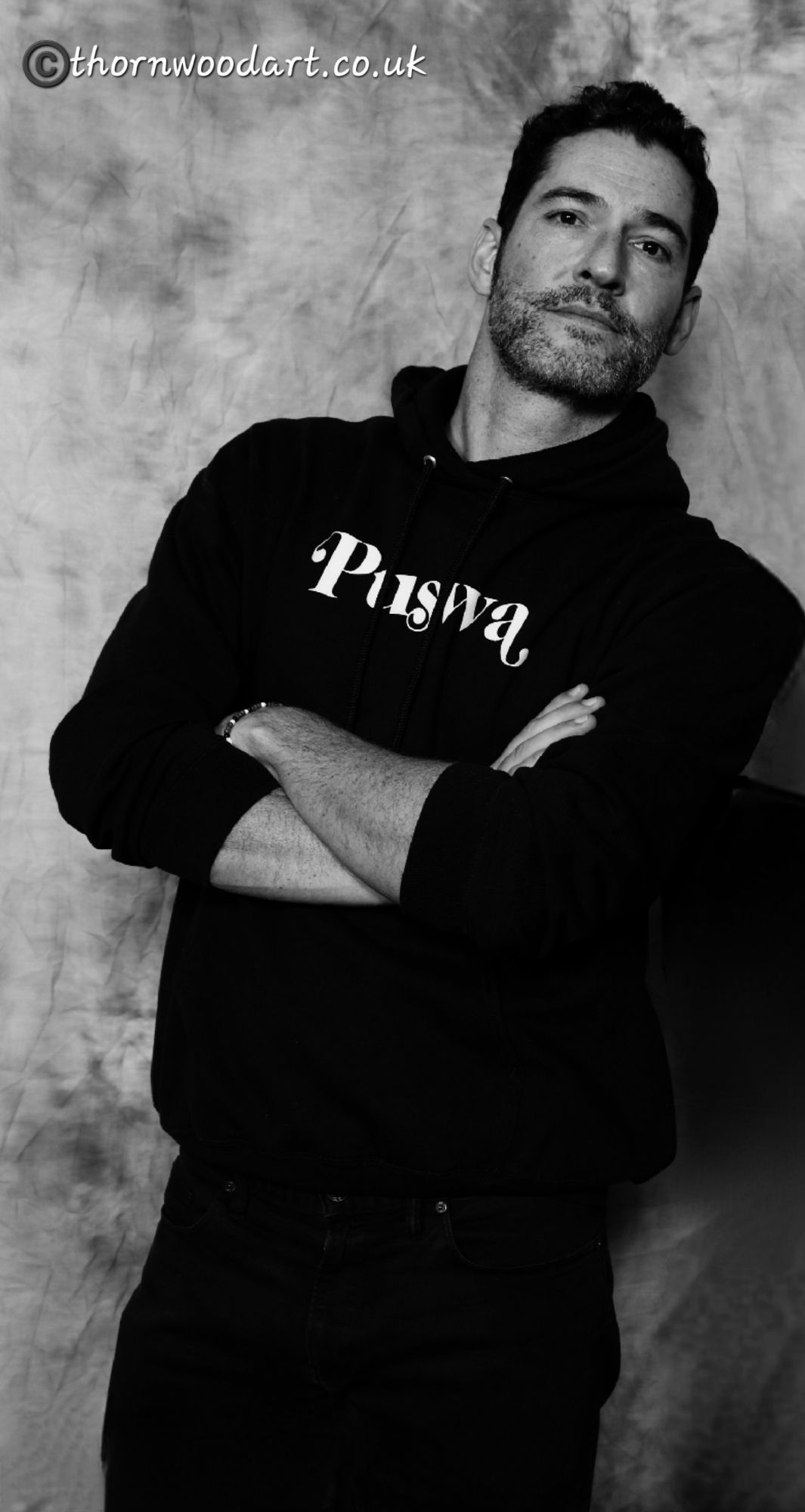 Tom Ellis, shown from head to thigh, leans casually to the right, arms crossed and looking at the viewer, neutral expression. He wears black jeans and a black hoodie with 'Puswa' written in white across it (from his wife's show 'Tell Me Lies').