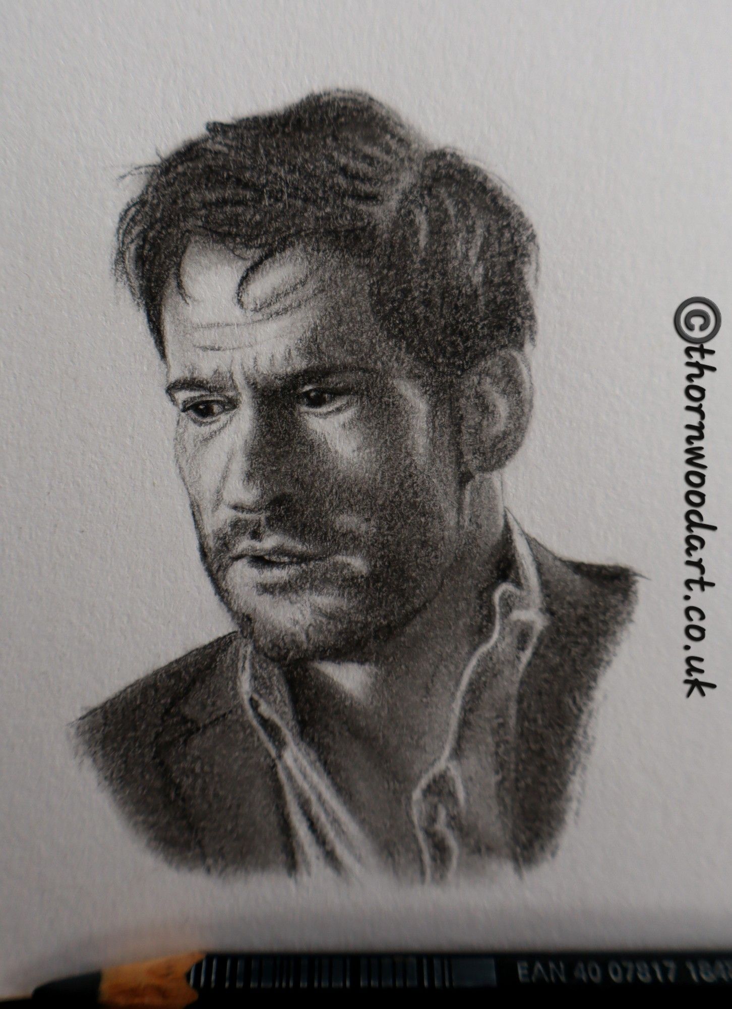 Close up photo of a very small but detailed pencil drawing of Tom Ellis as Lucifer Morningstar. A head and shoulders portrait, he faces the viewer but looks down to the left, dishevelled and sad. A pencil lies across the bottom of the drawing, and printed text states copyright Thornwood Art dot co uk.