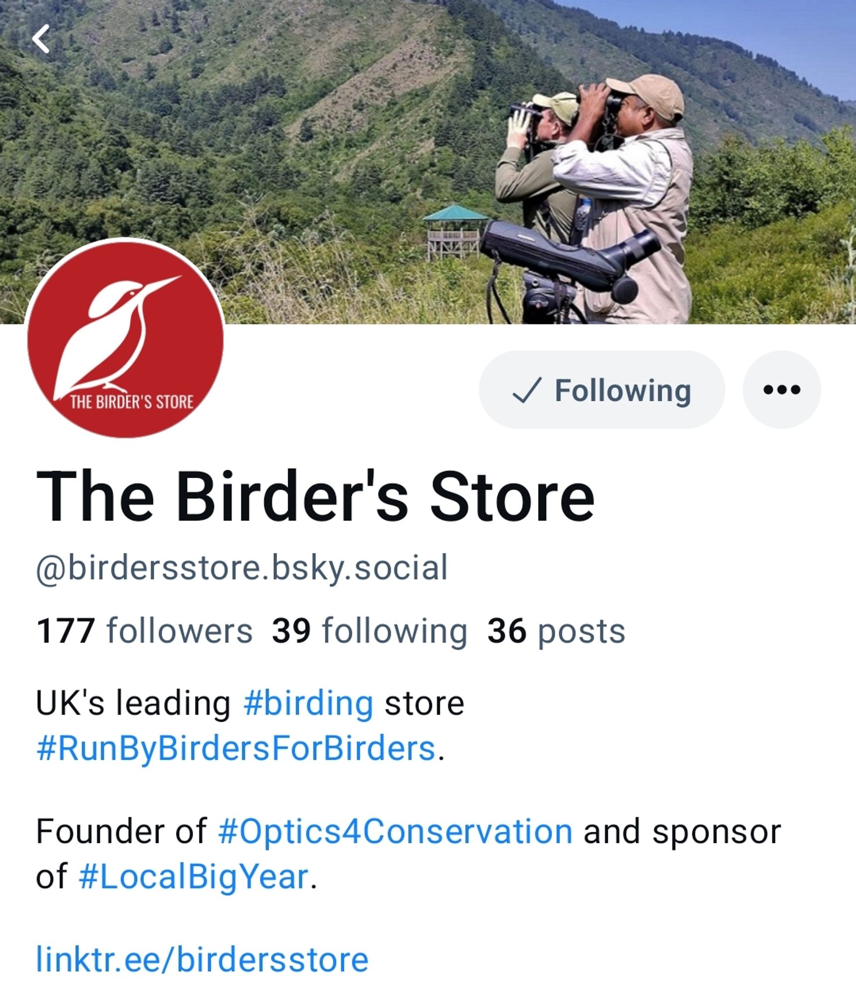 A screenshot of the Bluesky header for The Birder's Store with an image showing two birdwatchers looking through binoculars, with a spotting scope beside them, against a backdrop of green mountains.