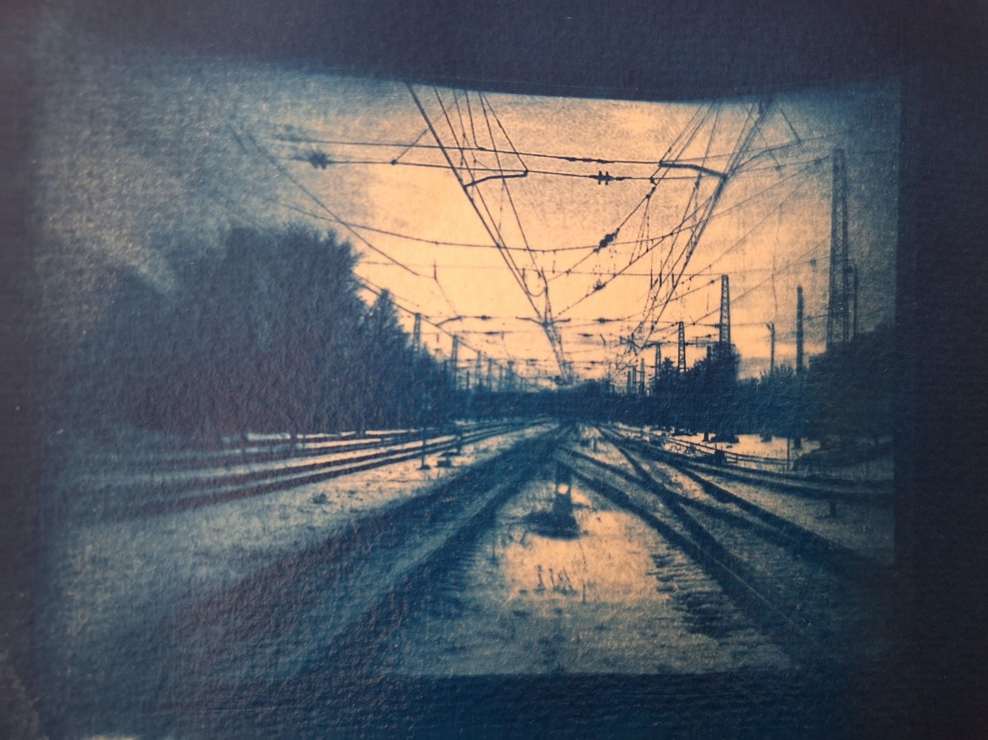 A blue-white monochrome photo of a railway track in winter. Edges are blurred and some texture of the paper is showing through the pigment