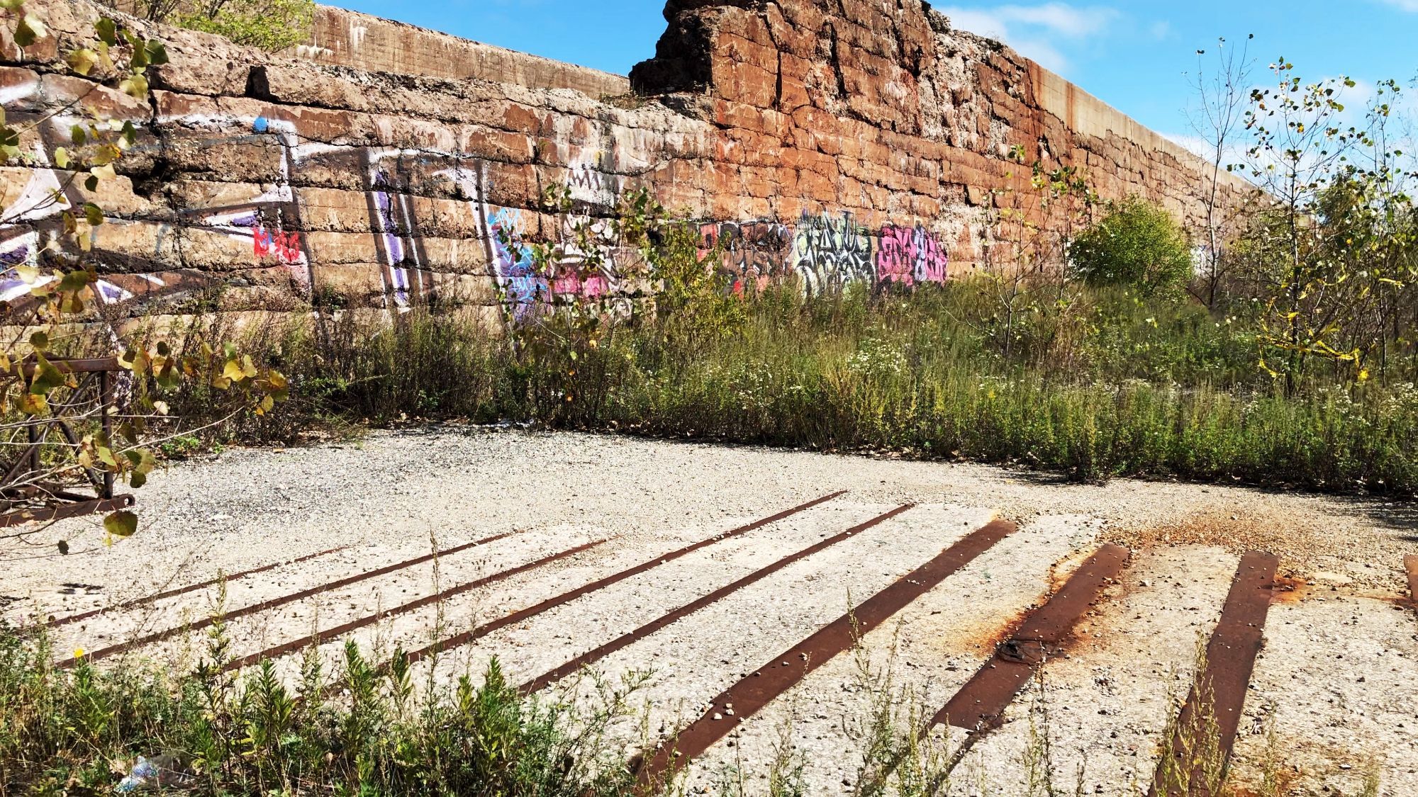 a still from a video showing a strange brick and stone ruin with graffiti, edging a concrete area of ground containing rusted strips of flat iron sort of like a washboard. beyond the paved part, a grassing-in and regrowing area stretches into the distance.