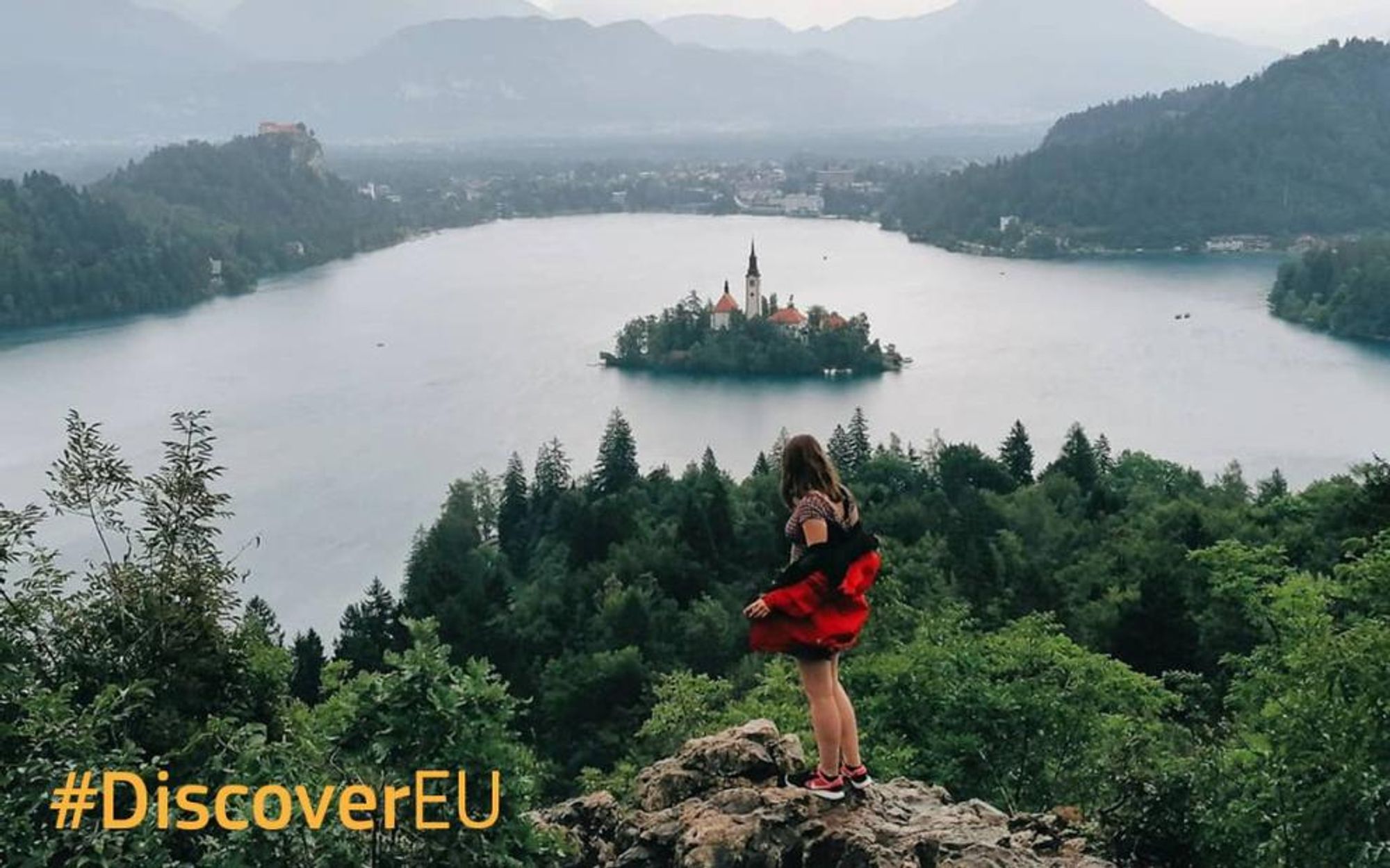 Blick auf den Bleder See in Slowenien, eine junge Frau steht dem Betrachter abgewandt hoch oben auf einem Felsen und schaut hinunter auf den See und die Insel mit der Marienkirche. Dazu der Hashtag #DiscoverEU