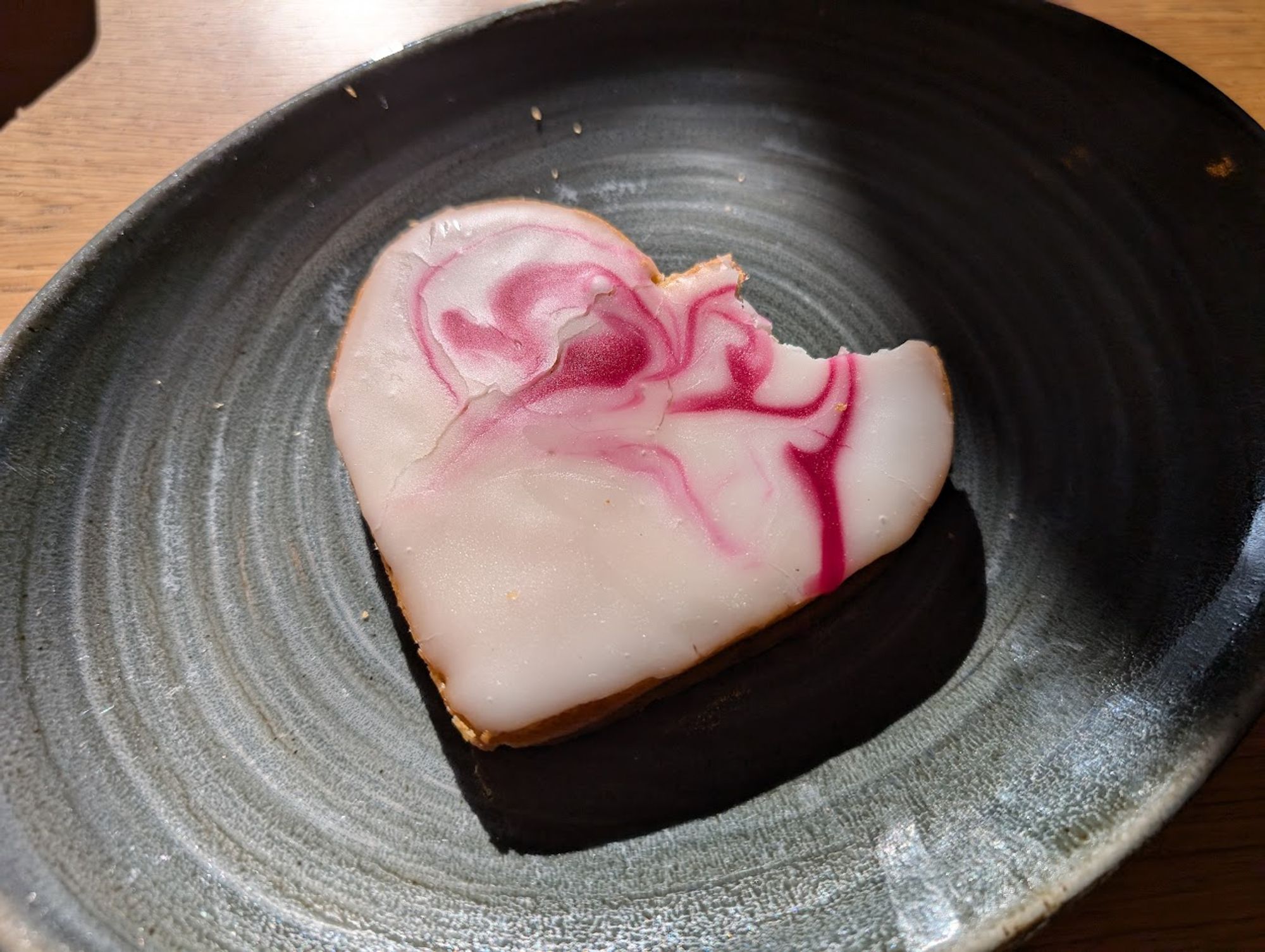 A piece of pastry, shaped like a heart, with white and pink red sugar coating, with a bite taken out of it.