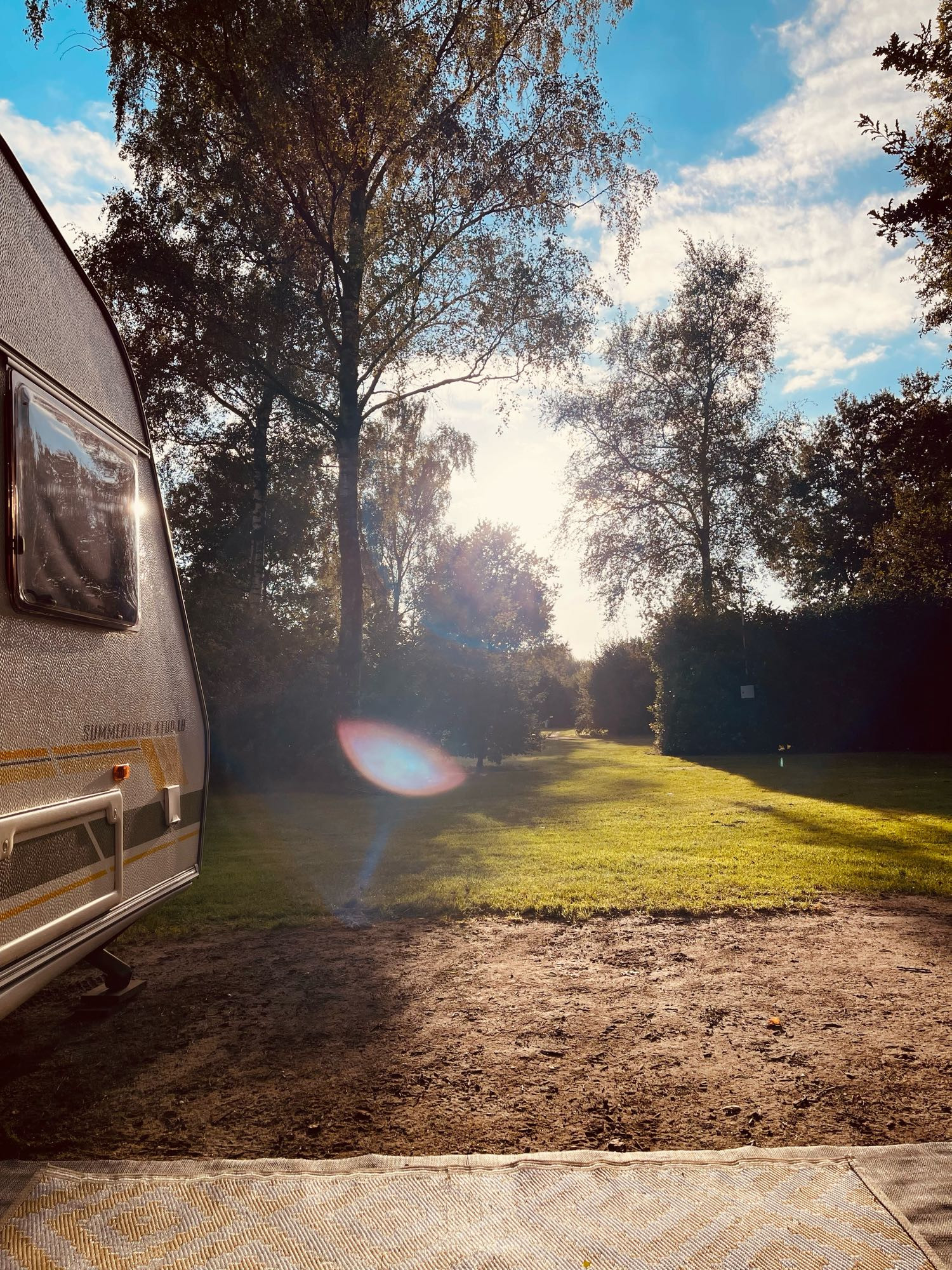 Foto met links een stukje van een caravan. Op de voorgrond en maar, zand en verder een graspad met heggen en bomen. De zon staat laag te schijnen