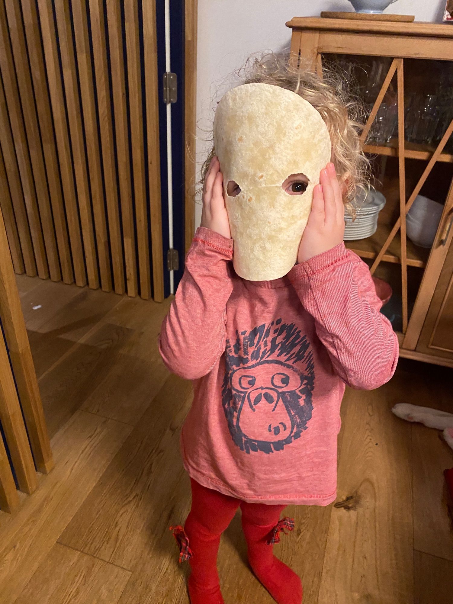 Foto de una niña de tres años vestida con leotardos rojos con un lacito y una camiseta tambié roja con una cara de gorila perplejo dibujada. A la niña no le vemos la cara porque se la tapa con una tortilla de trigo en la que ha mordisqueado sendos agujeros para los ojos. Unos rizos rubios asoman por encima de la tortilla. It’s giving el niño de El Orfanato, pero también es bastante adorable, la verdad, y no lo digo porque la haya parido yo.