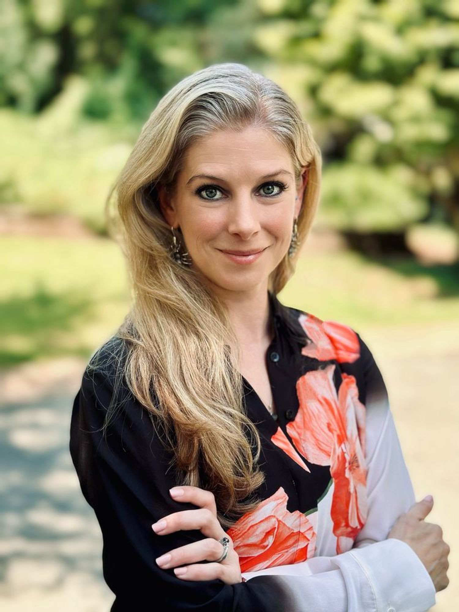 A professional photograph of Dr. Nadia Drake, a blonde women, smiling slightly at the camera, wearing a button down blouse that is black and white with a band of huge red flowers crossing from left shoulder down across her torso. Her arms are crossed in front of her, and there is a blurred background of trees and grass.