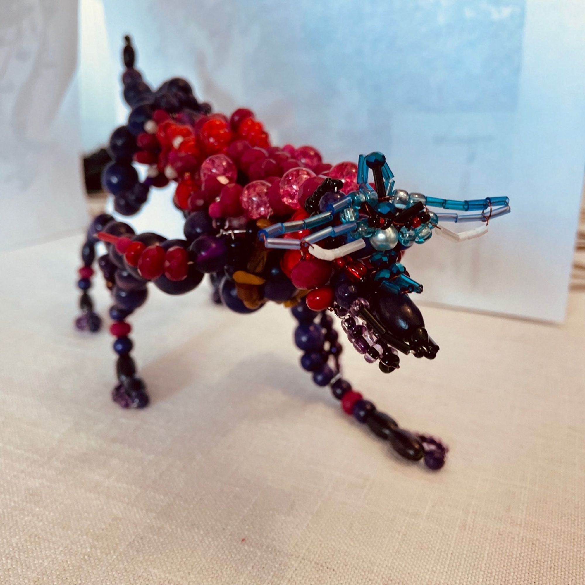 WIP sculpture of Oki (in wolf form) from Okami; posed in battle ready position facing viewer diagonally; purple, red and pink beads make up the bulk of his body; thin blue beads make part of Oki's mask (unfinished); his mane and sword are still missing.