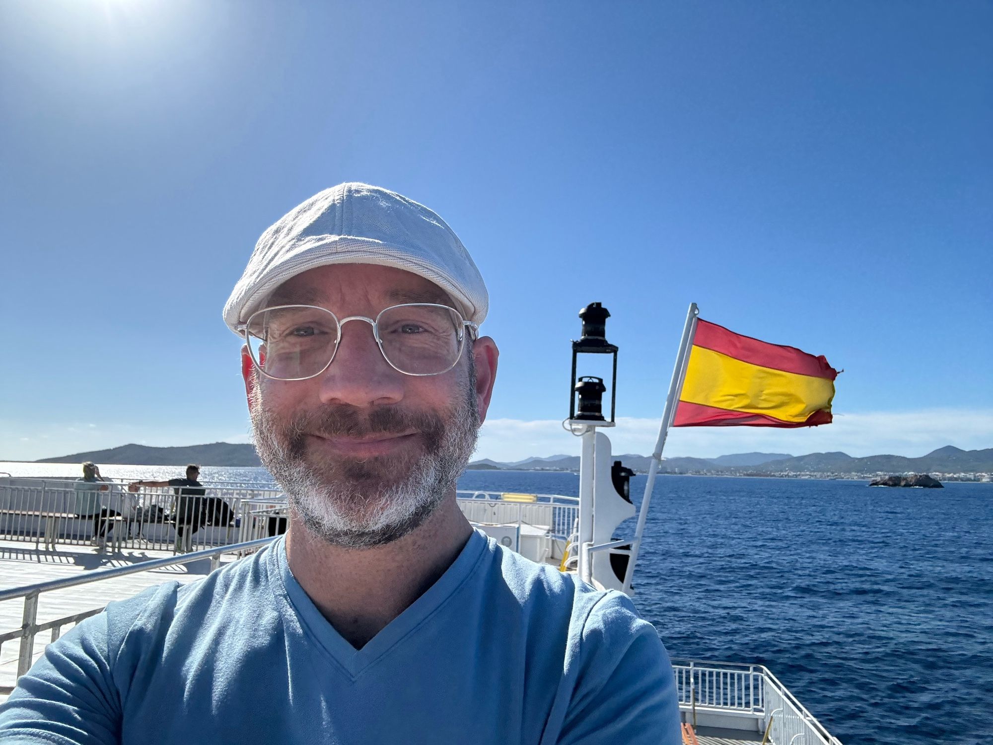 Selfie auf der Fähre von Ibiza nach Formentera. Im Hintergrund das Heckpositionslicht und die Spanische Flagge, dahinter ist die Küste von Ibiza zu sehen. Und alles unter blauem Himmel und auf blauem Meer.