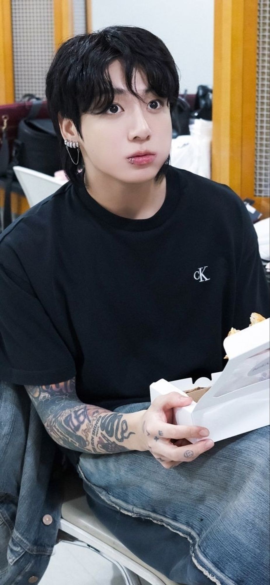 Jungkook in a black CK T-shirt and baggy jeans eating something from a white box. He's looking up at something like a dear in headlights, his eyes wide.