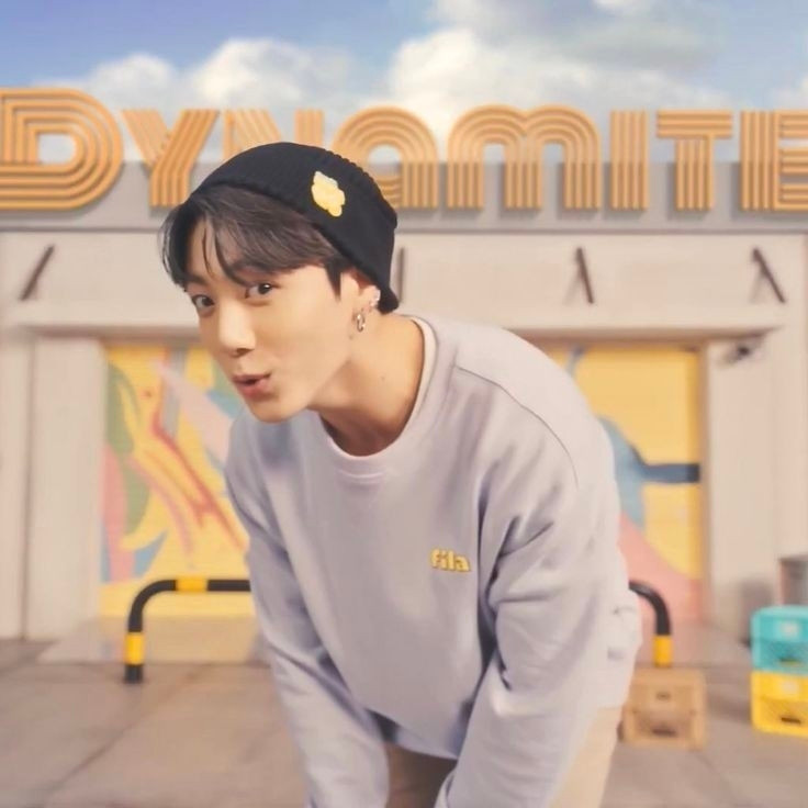 A happy Jungkook in front of a Dynamite sign wearing a lavender sweatshirt and black beanie from Fila.