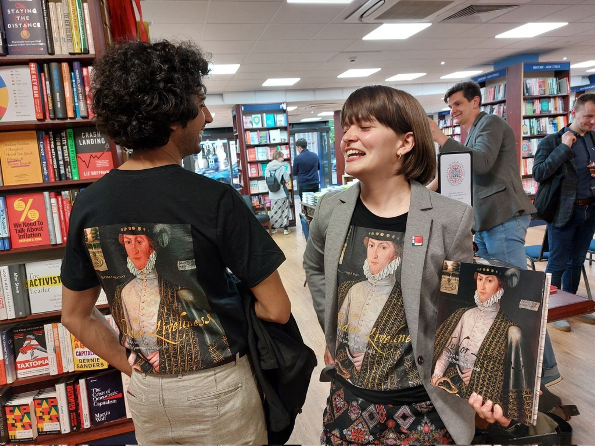 Two people standing in a bookshop. One is holding a copy of Tudor Liveliness: Vivid Art in Post-Reformation England. They are both wearing t-shirts with the book cover printed on them.