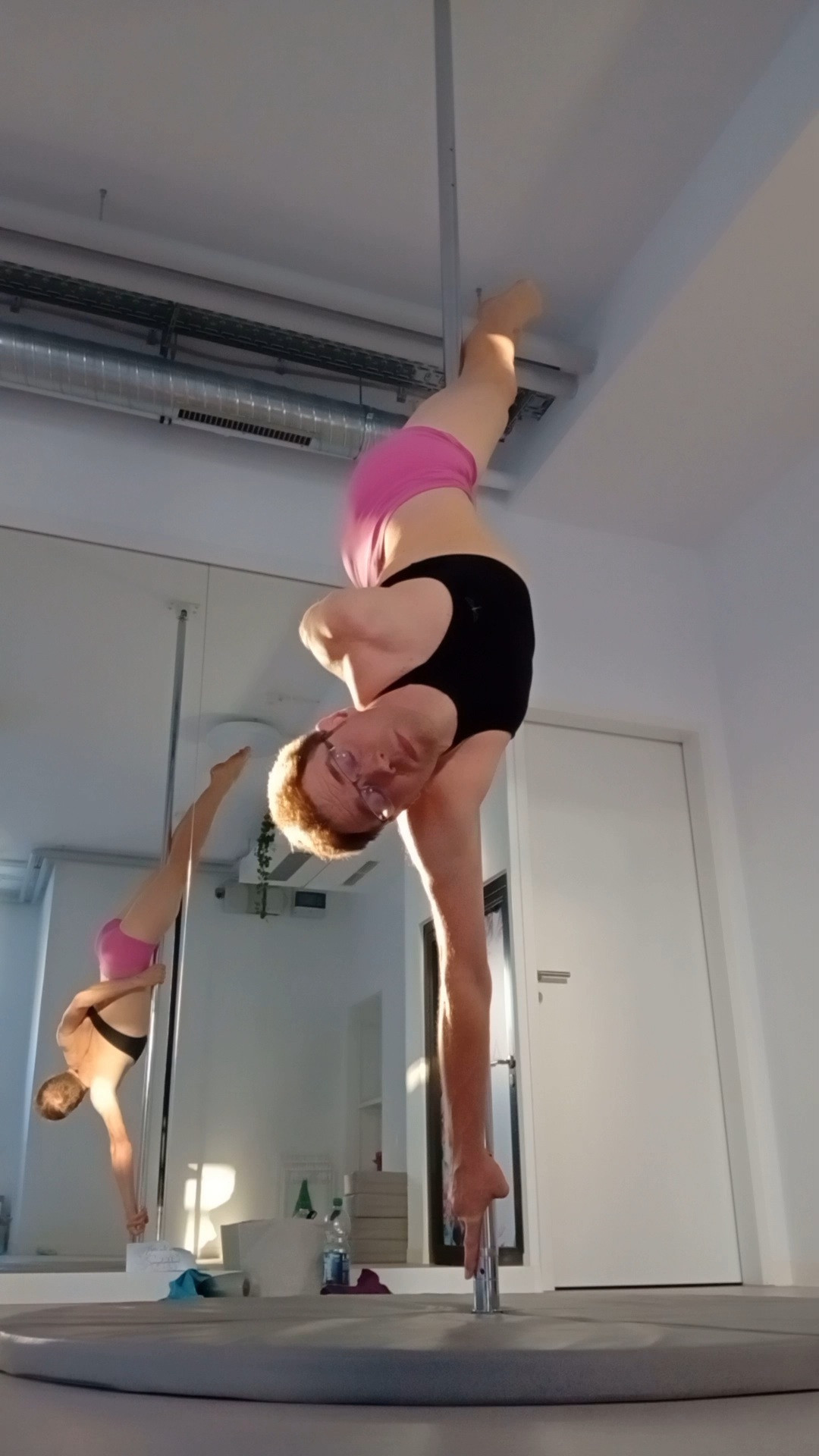 Me doing a pole shape in a pole studio. I'm hanging upside down on a chrome pole. The pole is between my knees, my inner hand is streched put above my head and holding on to the pole and my outer hand is holding on to the pole behind my back.
My body is almost straight and diagonal to the pole.
Behind me is a reflection of the shape in a large mirror.
I'm wearing a black nackholder sports bra and pink shorts