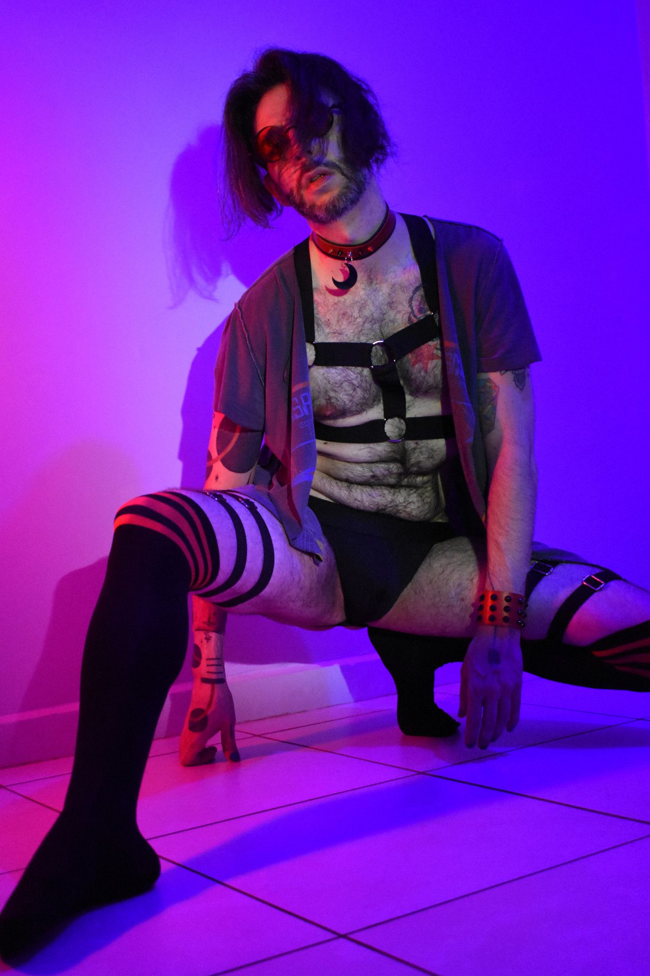 flux posing crouched on the floor with one leg flexed and the other extended. He's wearing round red sunglasses, a black harness, black thigh-high socks with some red stripes on the top part of it, a red choker with a black moon pendant on it, a red bracelet with black spikes on it, and a loose gray open t-shirt. The lighting in the room consists mostly of purple and red tones. flux is looking directly at the camera, a slight daring energy coming from his expression.