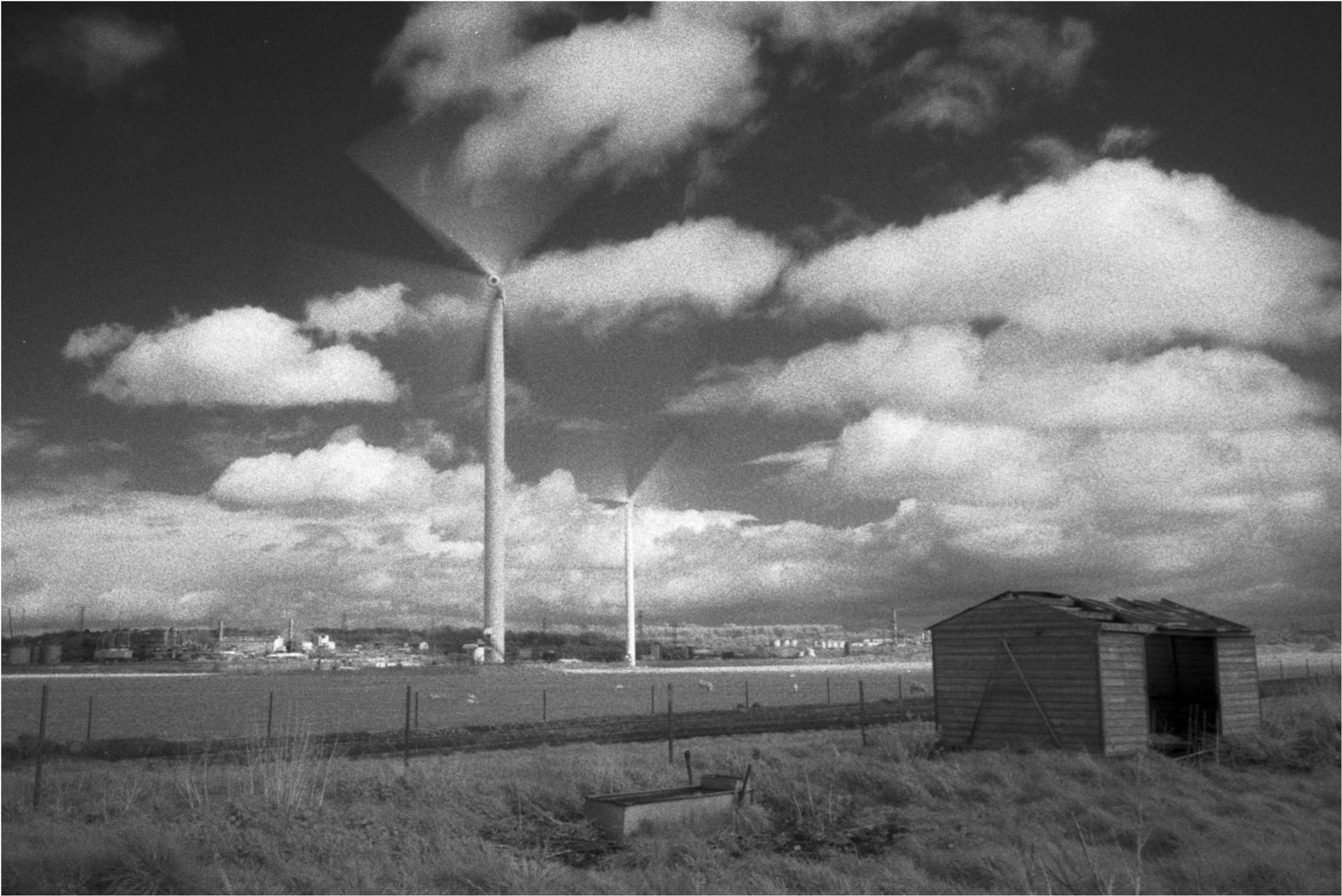 A high contrast monochrome landscape photograph. There is a shed in the foreground, wind turbines in the midground and a chemical plant in the background.