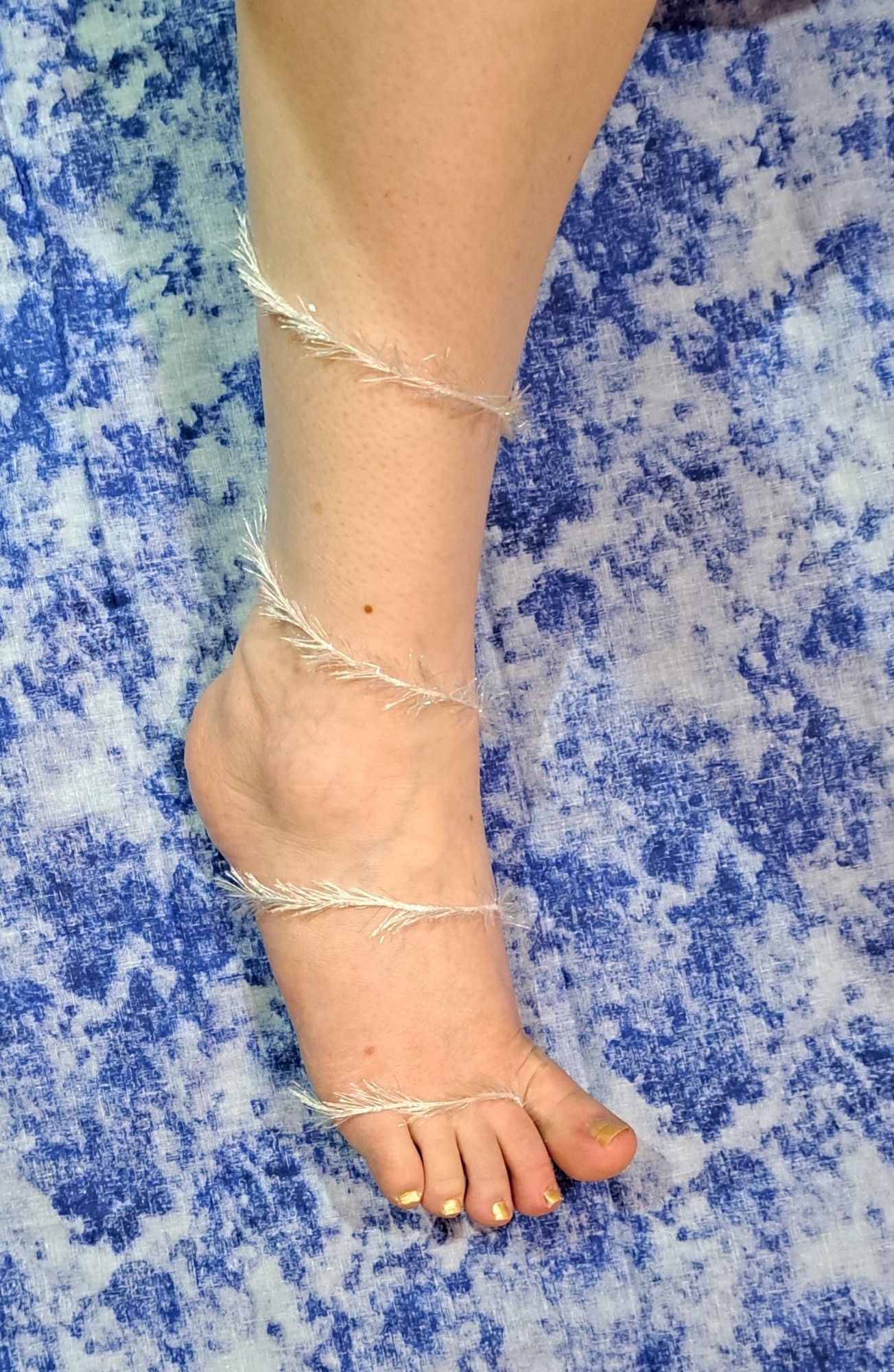 An iridescent white feathery ribbon is wrapped around her lower leg, ankle,  and foot. Gold nail polish.