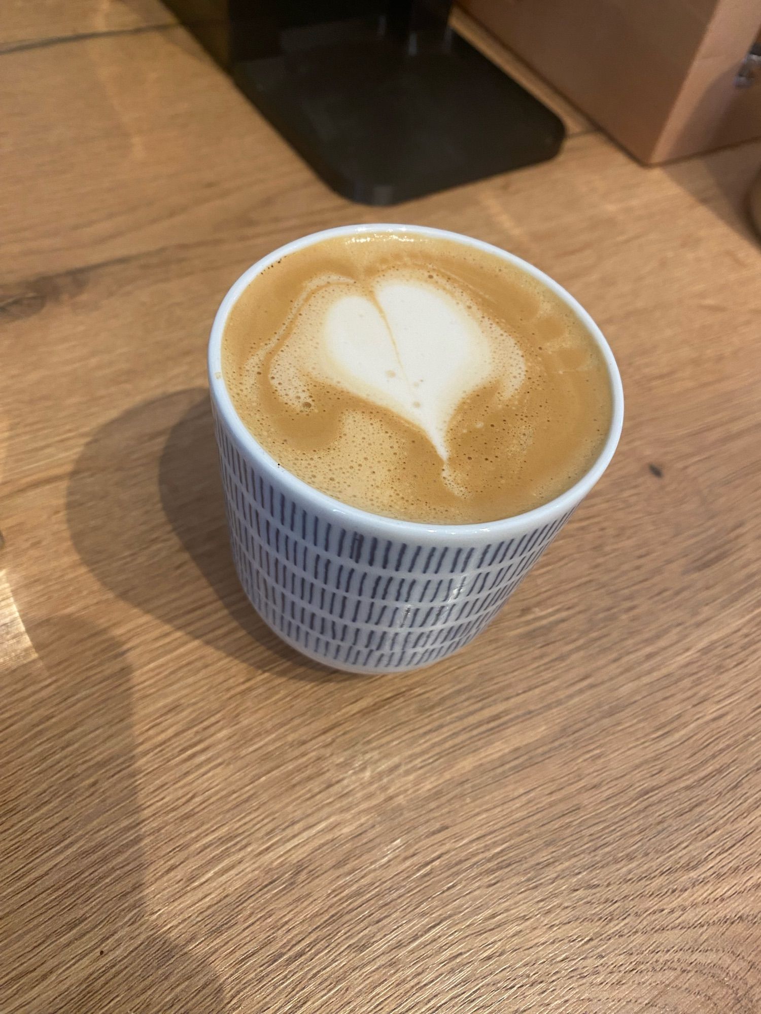 Flat white in blau weiß gestreiftem Becher. Herzchen mit Haaren Latteart inklusive. Platziert auf Holz Arbeitsplatte.