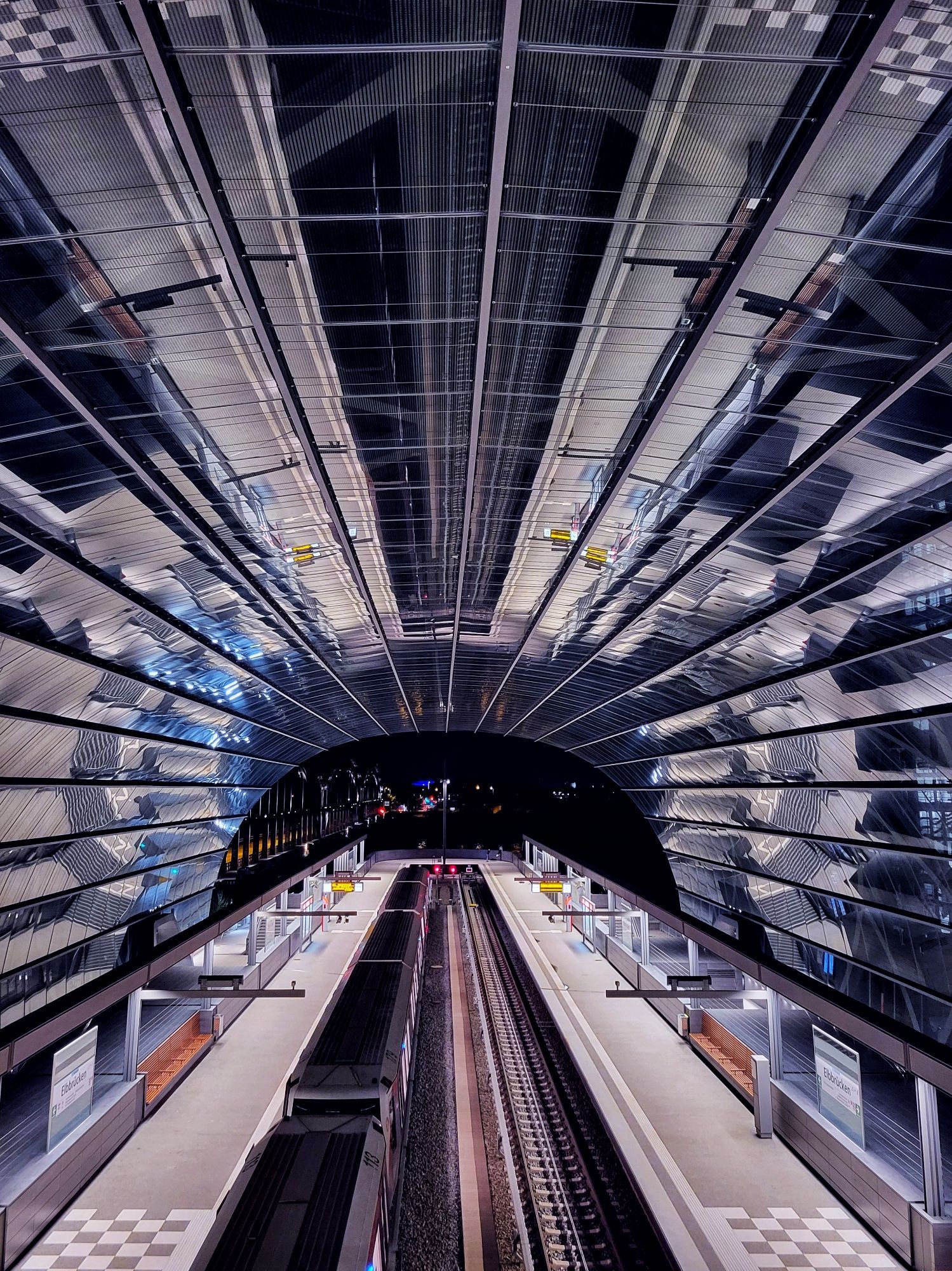 Blick von erhöhter Position. Unten sind Gleise. Links steht eine U-Bahn. Die Station ist überdacht. Die Überdachung ist aus Metall und Glas. Die Linien führen nach hinten. Hinter der Station ist ein Fluss, links ist eine Brücke zu erahnen. Es ist dunkel. Überall sind kleine Lichtpunkte. Nur die u-Bahn-Station ist hell.