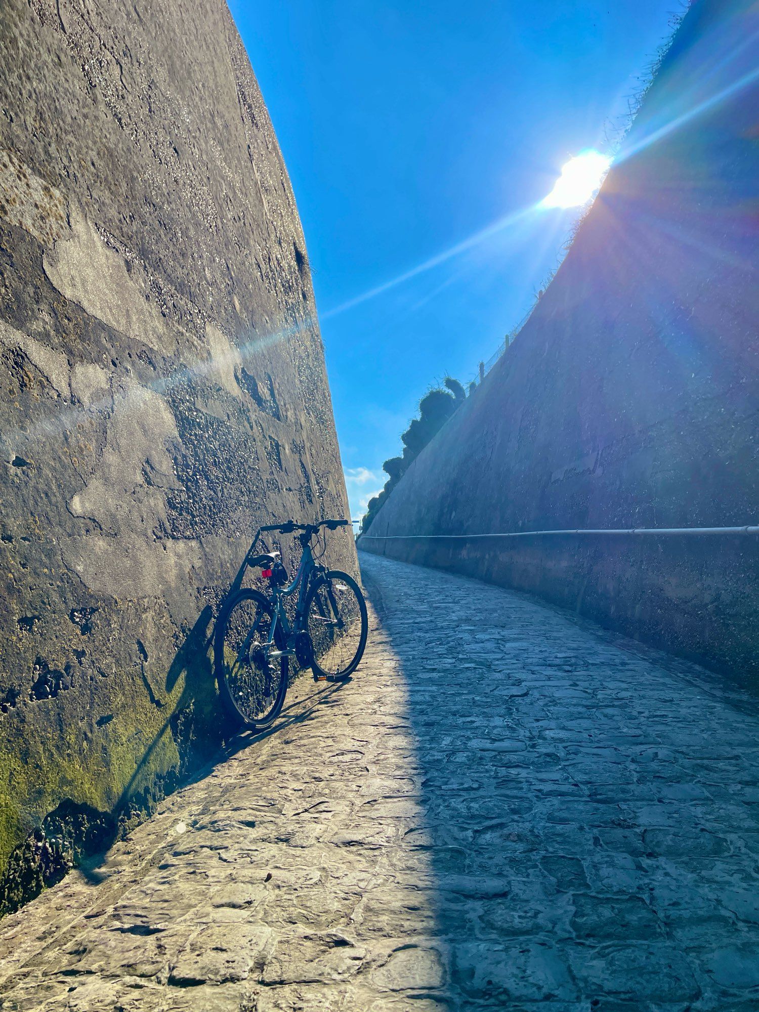 Bike leaning on the sunny side of a stone wall with a stone walkway leading up the hill