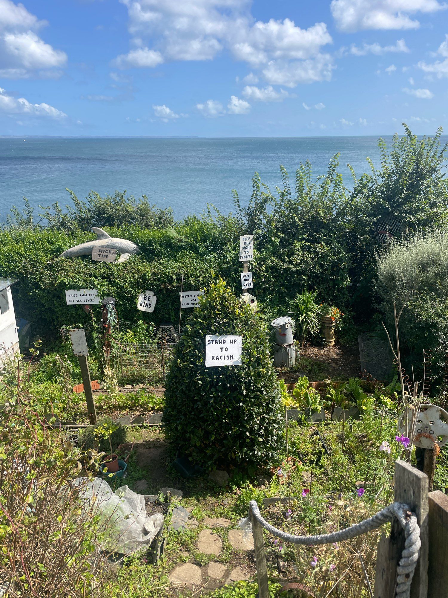 Community garden with amazing sea views and blue skies in Mousehole with handcrafted sculpture and trees bearing signs reading STAND UP TO RACISM, BE KIND, RAISE AWARENESS NOT SEA LEVELS and more inspiring activist statements