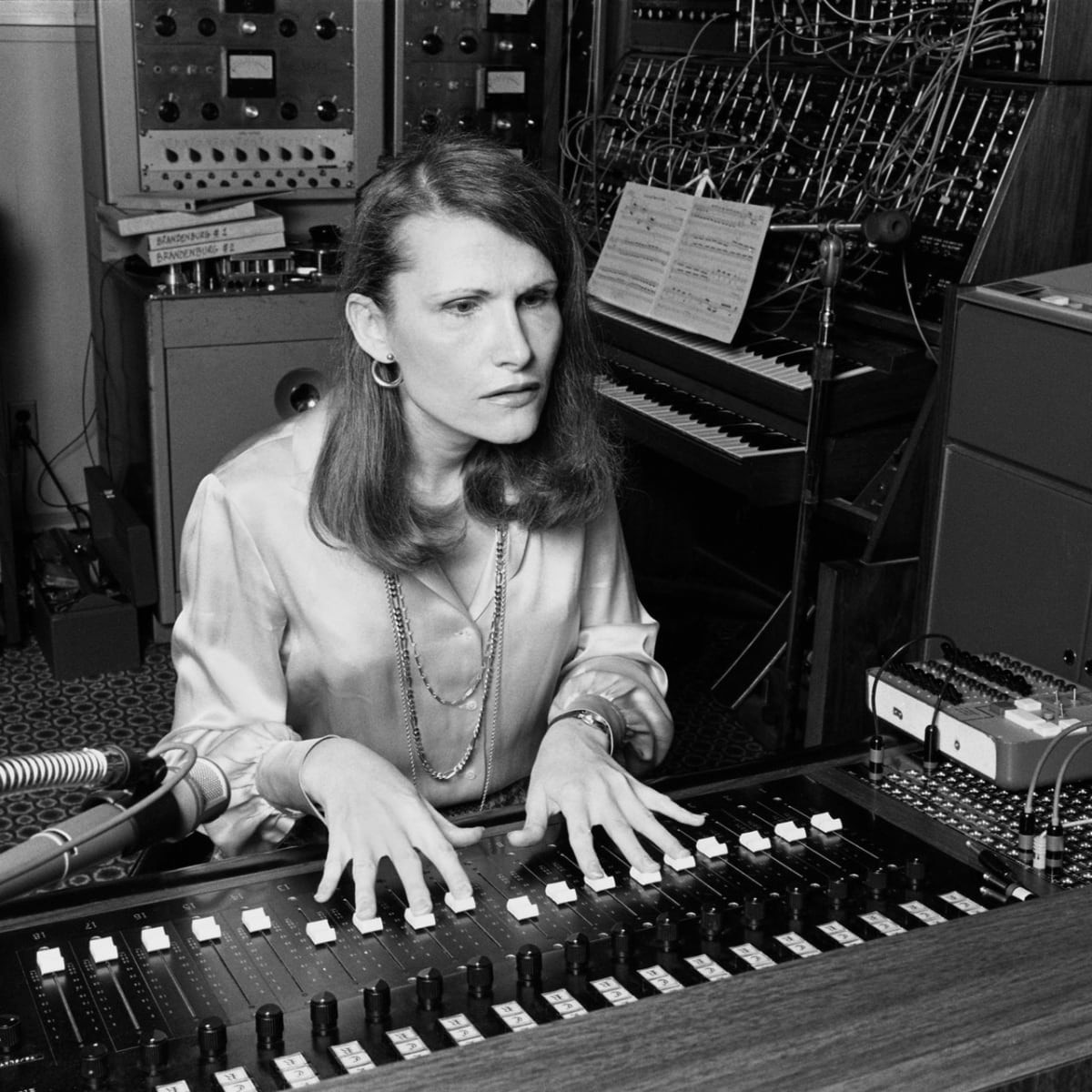 Black and white photo of Wendy Carlos, a woman with long, dark hair, pushing up faders on a mixing board with her fingertips. Modular Moog synthesizer and other studio equipment in the background.