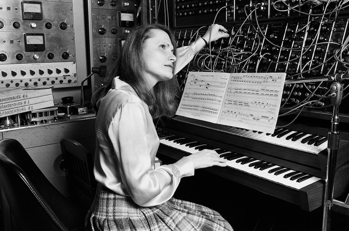 Black and white photo of Wendy Carlos, a woman with long, dark hair, seated at the keyboards of a modular Moog synthesizer. The synth is covered in 1/4" patch cables. Other studio equipment behind her.