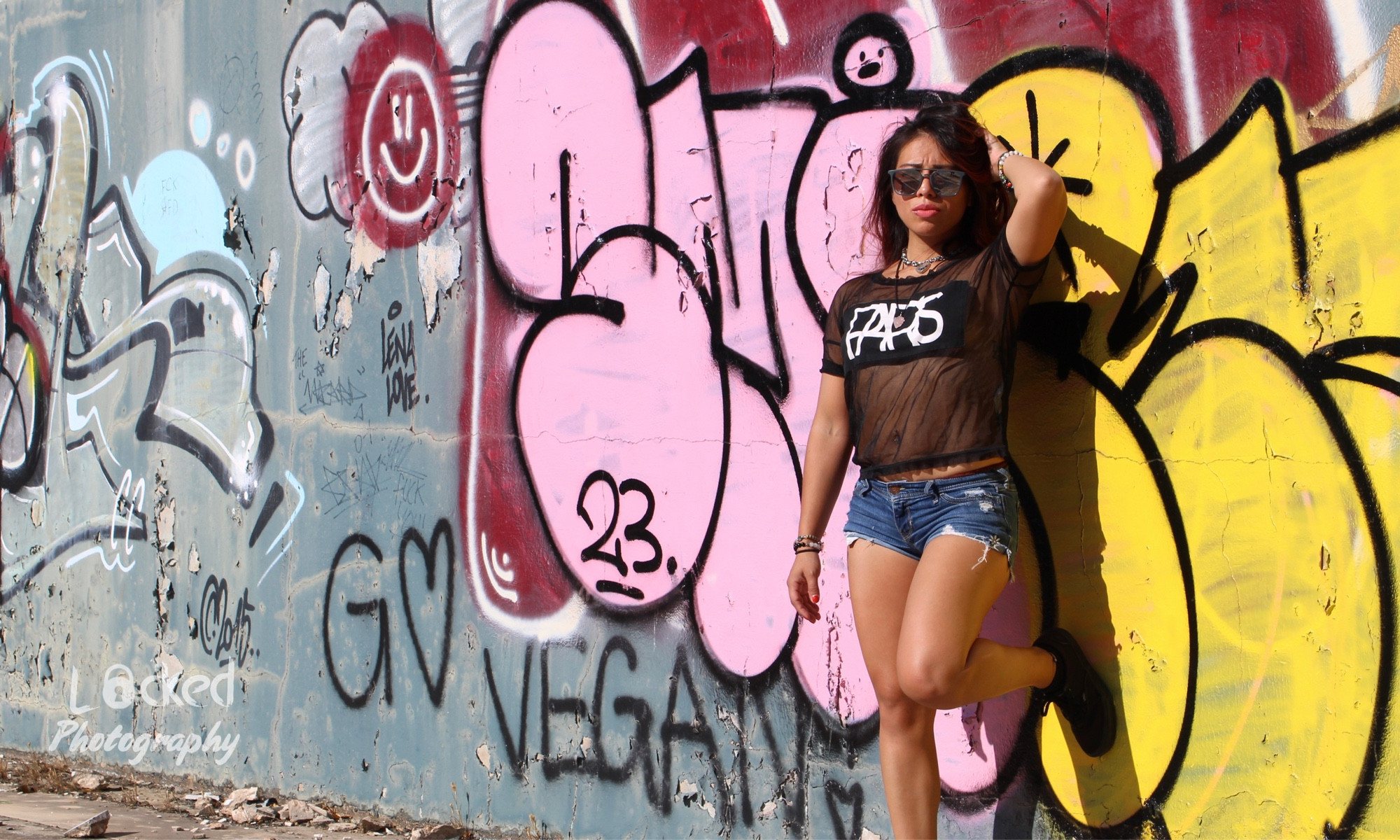 A latin girl wearing sunglasses, a mesh shirt and jean shorts poses against some colourful graffiti art