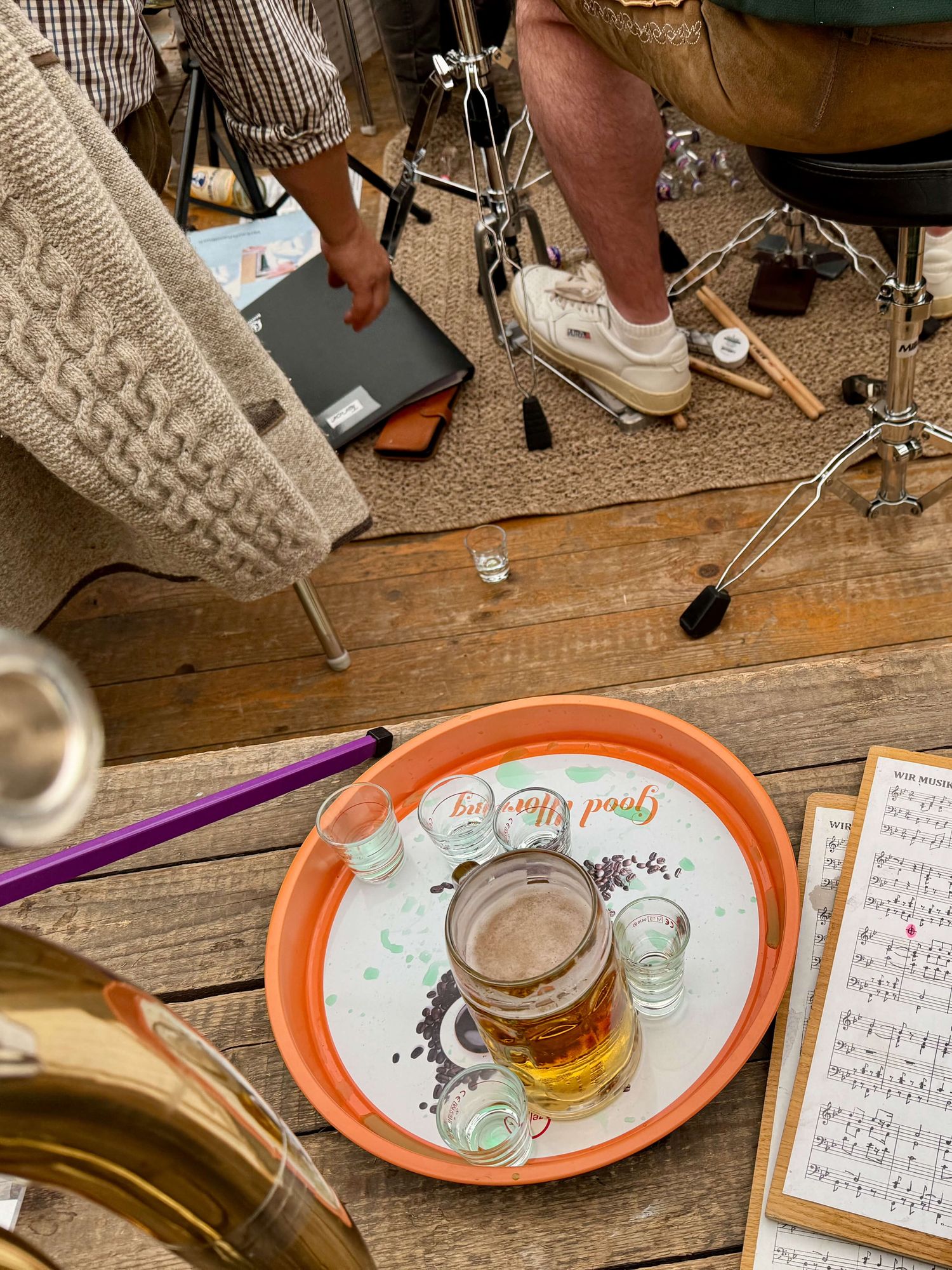 Auf dem Bild ist eine Szene von einem Liveauftritt während eines Frühschoppens zu sehen. In der Mitte des Bildes befindet sich ein orangefarbener Tablett, auf dem ein großer Bierkrug mit Bier und mehrere leere Schnapsgläser stehen. Das Tablett liegt auf einem Holztisch.

Rechts unten im Bild sind Notenblätter auf einem Klemmbrett zu sehen, was darauf hinweist, dass hier Musiker tätig sind und Live-Musik gespielt wird. Am unteren Rand des Bildes ist ein Teil eines goldfarbenen Musikinstruments, wahrscheinlich eine Tuba oder ein ähnliches Blechblasinstrument, zu erkennen.

Links im Bild ist ein Stuhl mit einer hellbraunen, gestrickten Decke darauf. Ein Mann, der ein kariertes Hemd trägt, beugt sich nach vorne und hält etwas in der Hand, das wie ein Notebook aussieht. In der Mitte des Bildes ist eine Person teilweise sichtbar, die auf einem Schlagzeughocker sitzt und helle Sneakers trägt. Der Boden besteht aus Holzdielen und einem Teppich, auf dem Schlagzeug-Hardware und Drumsticks liegen.

Die Szene wirkt etwas unaufgeräumt und gemütlich, was auf die entspannte und lockere Atmosphäre eines Frühschoppens hindeutet.