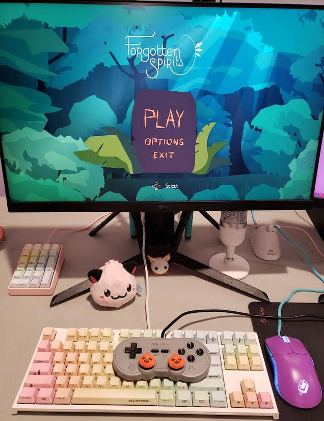 A picture of my monitor + PC setup. With the game Forgotten Spirits on the screen. Also a cat-poring plushy, a Dango figure, a keyboard with rainbow keycaps, a controller with jack o lantern thumbsticks, and other peripherals.