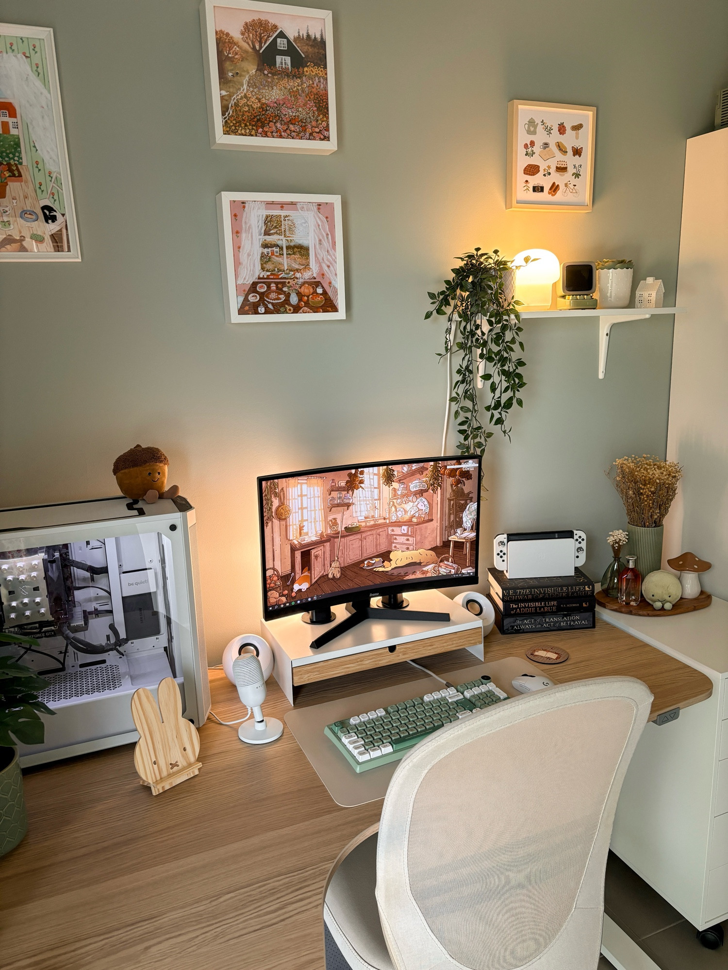 a photo of lili’s cosy desk set-up. wooden desk, white chair, a screen with a brown illustrated wallpaper, a green keyboard, a white pc, a white nintendo switch on top of stacked books, plants, and accessories in earthy or green colours. 