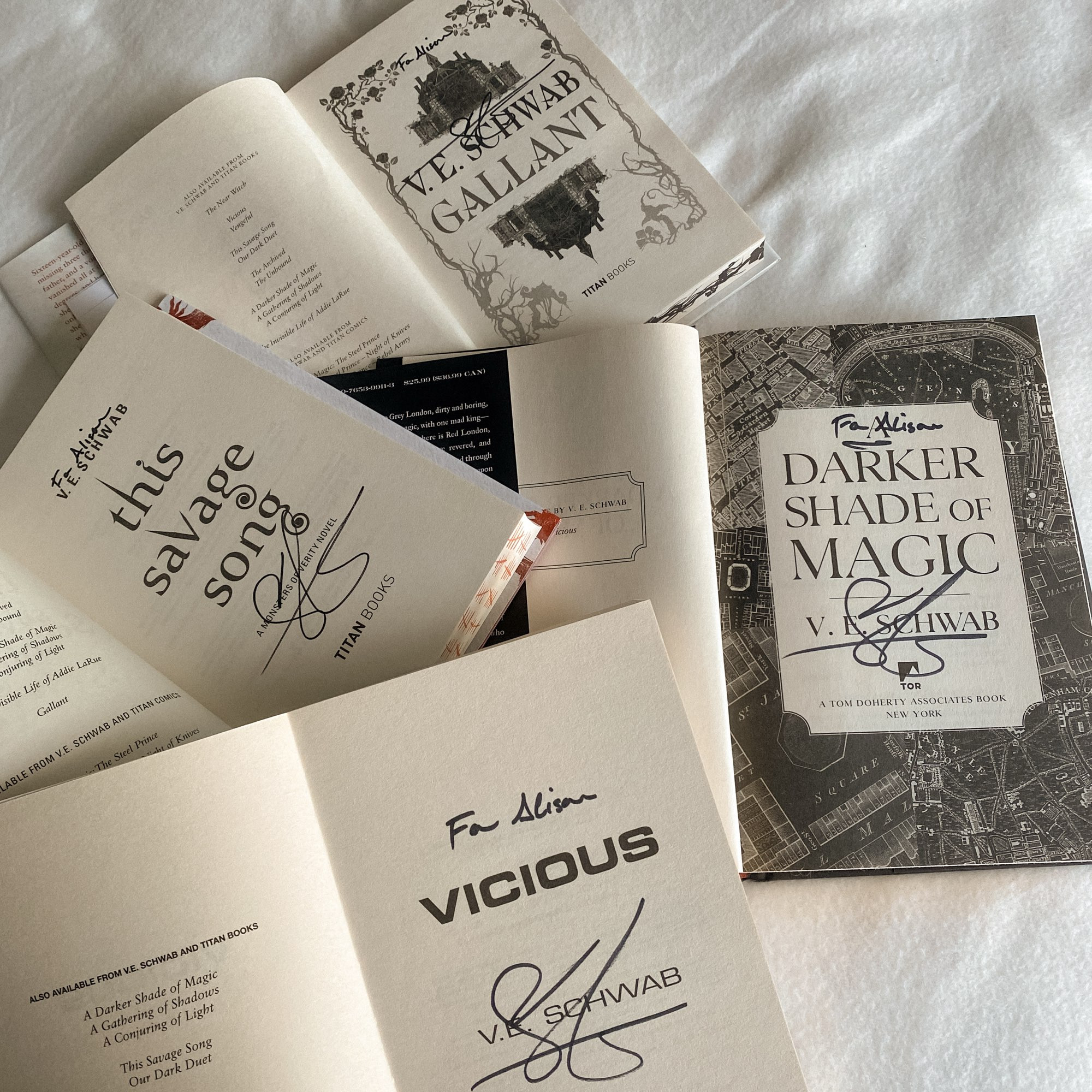 a white bedsheet with four v.e. schwab books laying disorganized on top. they are opened to the title page and are all signed and personalized to alison: vicious, a darker shade of magic, gallant, and this savage song. 