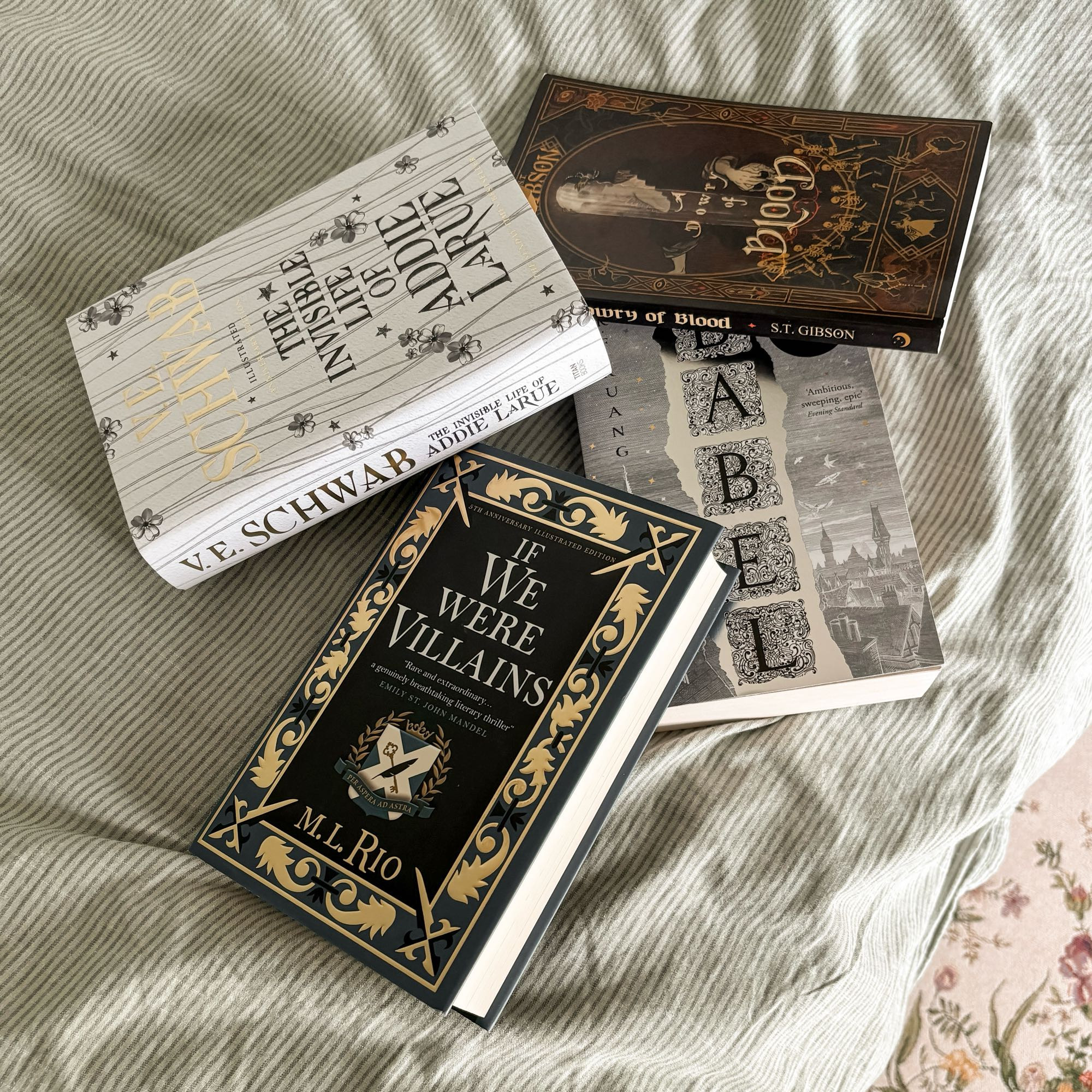 green and beige lined bedsheet with 4 books laying disorganized on top: the invisible life of addie larue by v.e. schwab, if we were villains by m.l. rio, babel by r.f. kuang, and a dowry of blood by s.t. gibson 