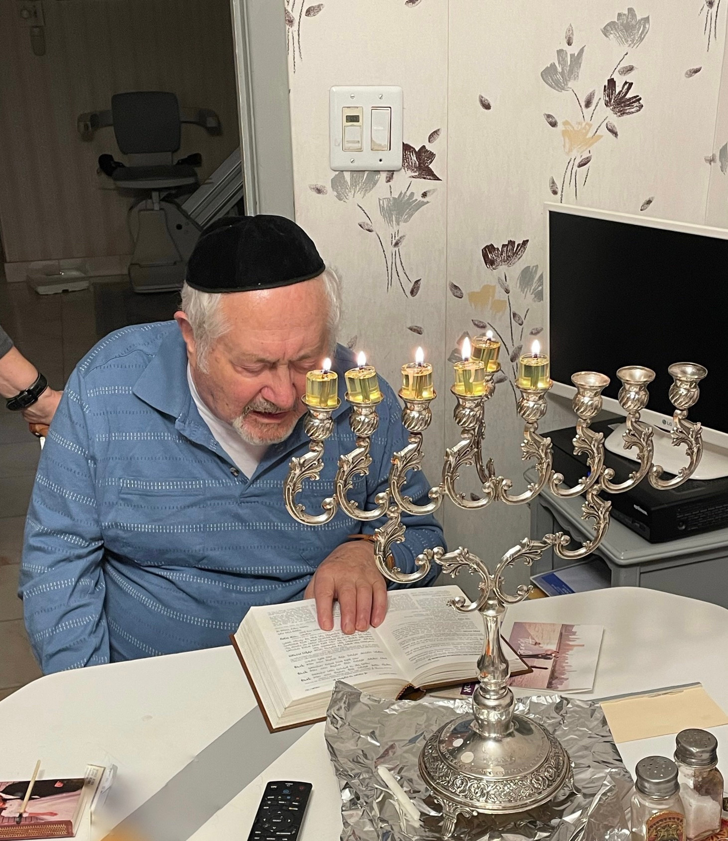 a 96-year-old man reading from a hebrew prayer book while lighting the hanukkah candles