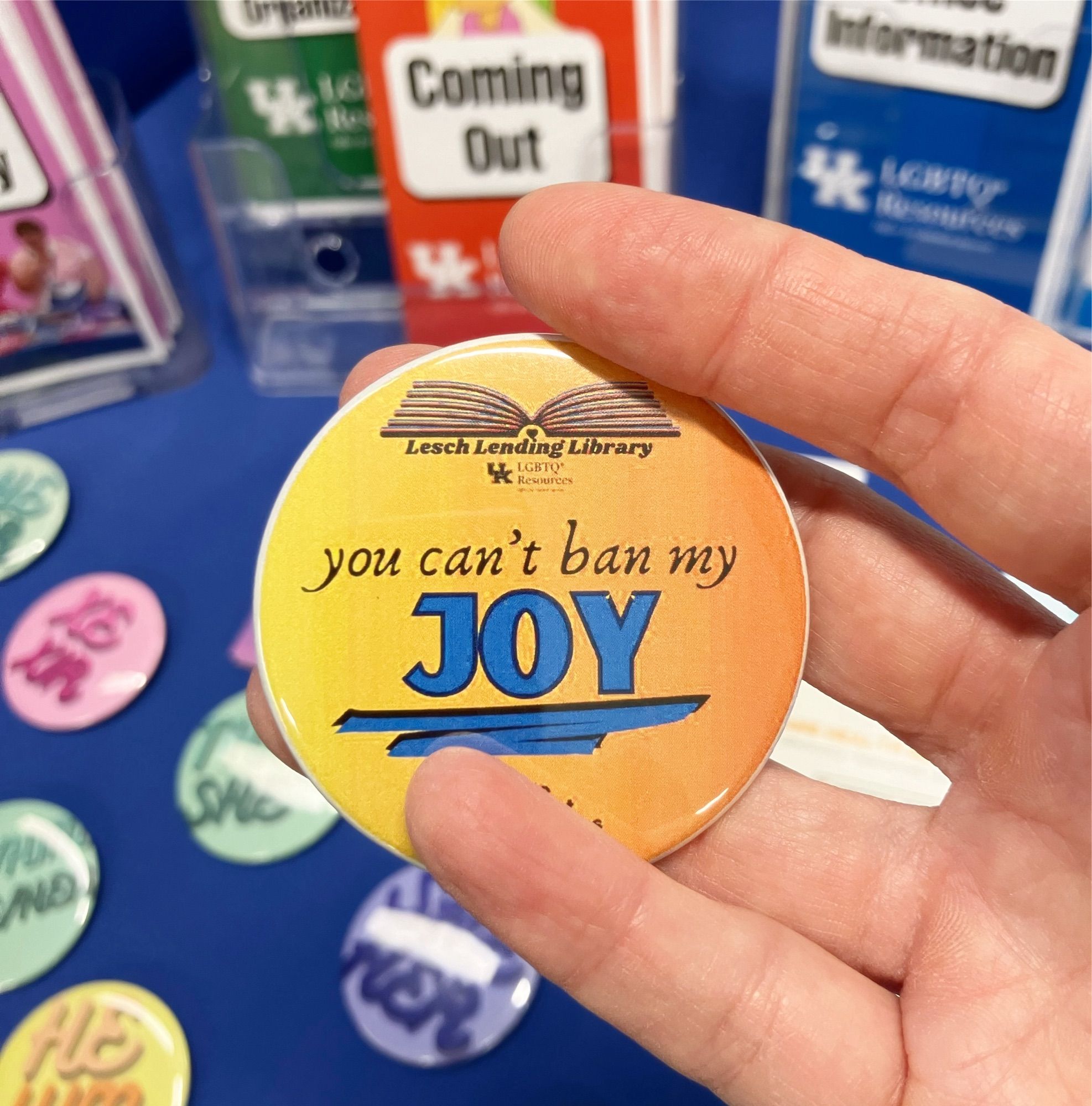 photo of a hand holding a button with logo is the lesch lending library and uk that reads “you can’t ban my JOY”. blurry in the background are pronoun buttons and pamphlets about coming out.