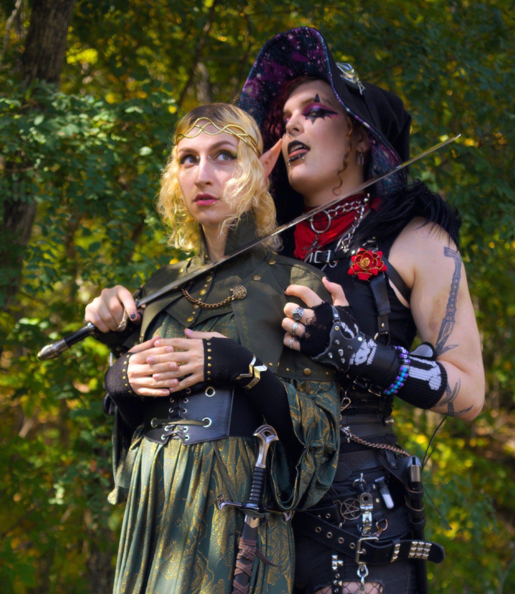 same girl, this time from the front, with a tall, strong goth babe behind her wearing a black outfit with lots of buckles and straps and chains and a witch hat, gripping her shoulder and holding a sword to her neck.