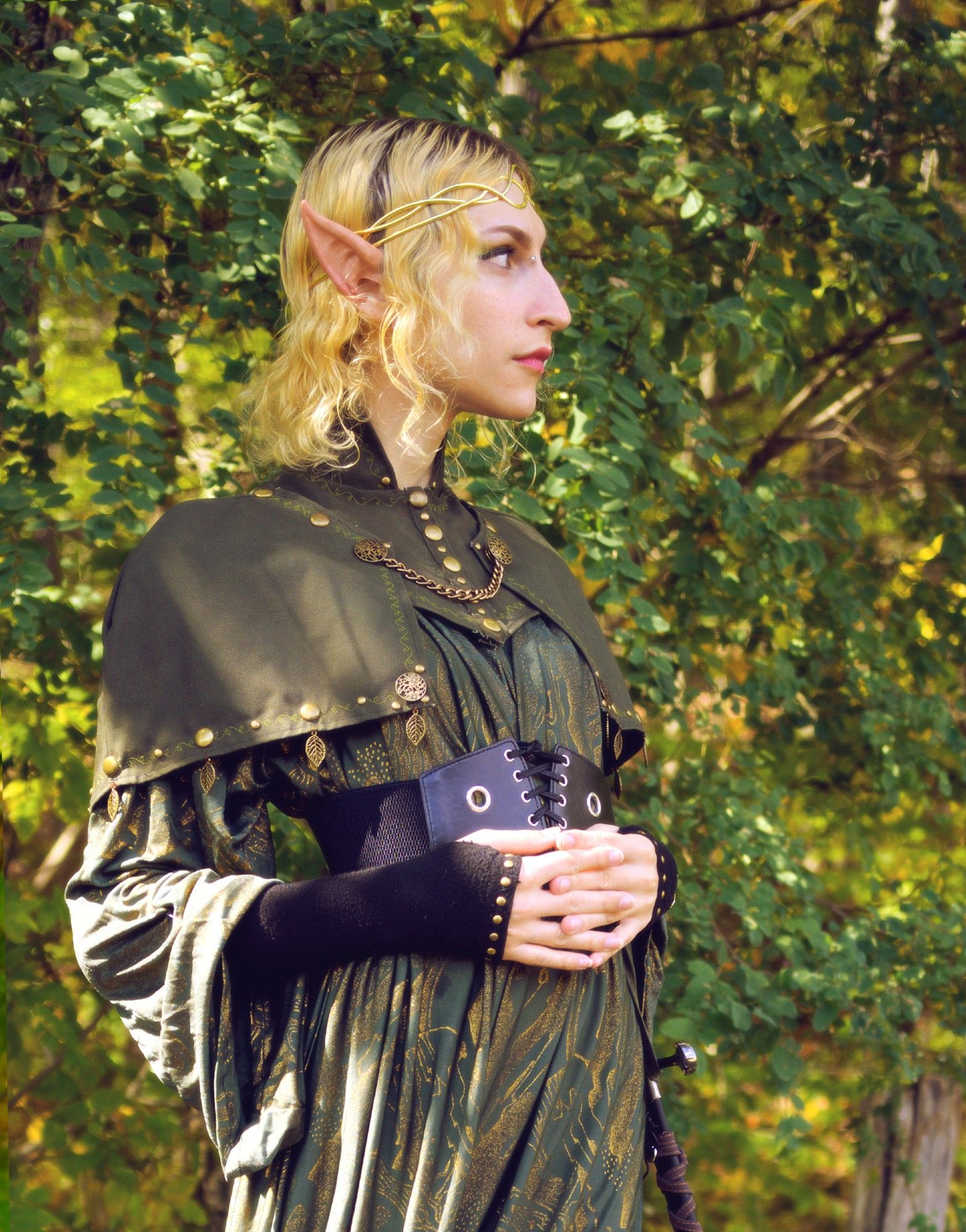 photgraph, waist up, profile. slim white tgirl with blonde hair wearing long elf ears and a green wizardly dress and green capelet with a high collar and brass adornments, and a gold tiara.