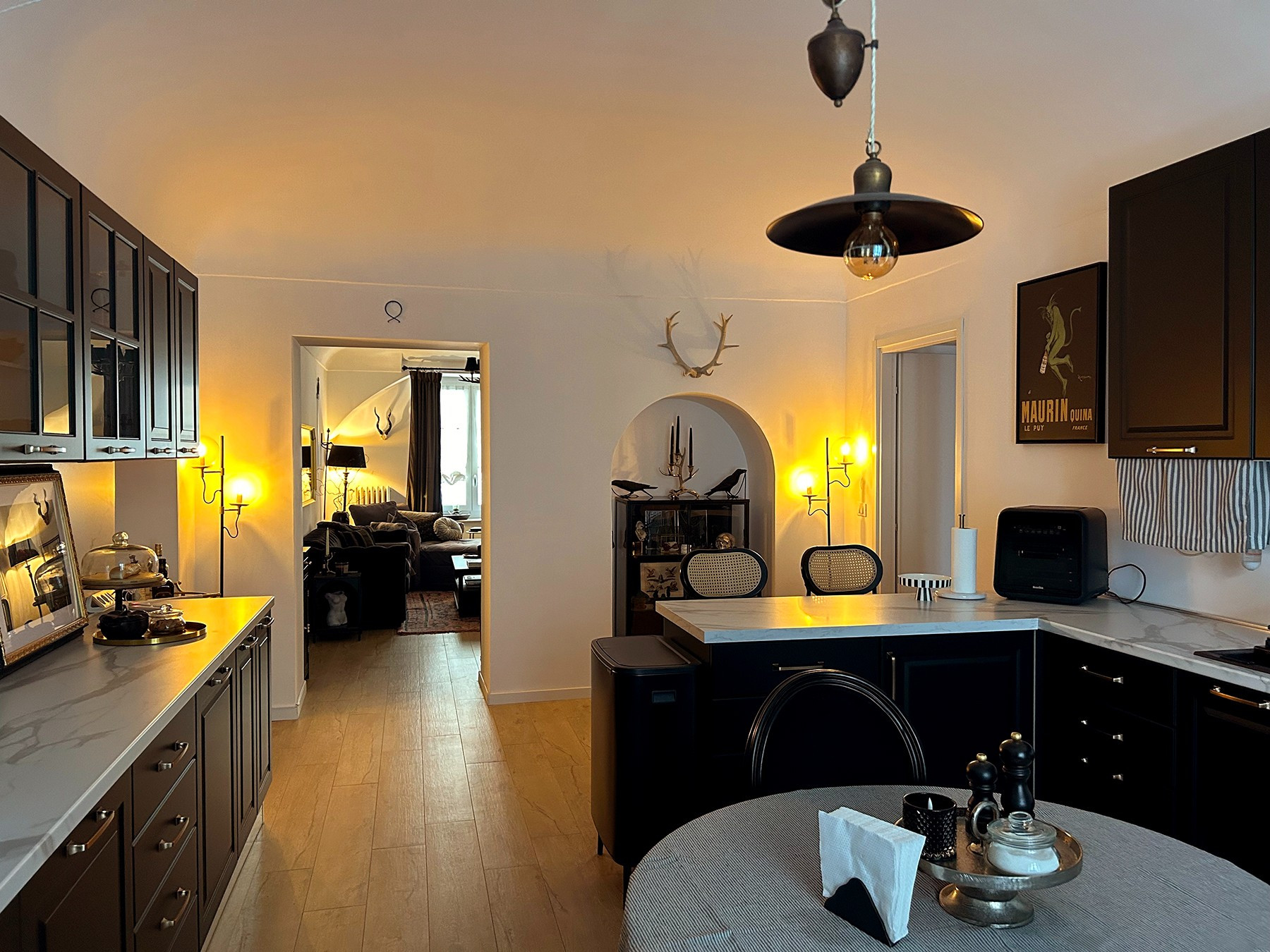 photo of an Italian apartment with a small kitchen, walkway, and doorway leading into the living room. The kitchen cabinets are black with brass hardware, the countertops are faux marble, and a brass pendant lamp hangs from the ceiling over a round Ikea dining table.