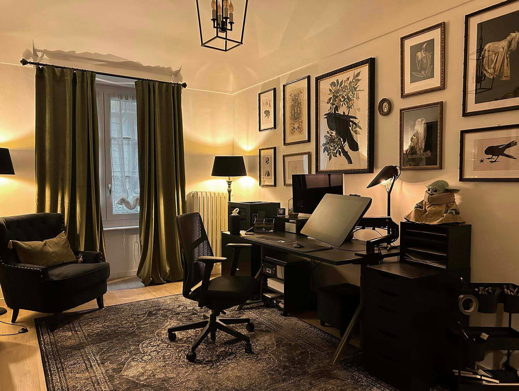 photo of a small studio room in an Italian apartment. There's an area rug, a black Ikea work desk with a cintiq and computer, and a black Ikea chest of drawers. On the opposite wall is a black arm chair. The window has olive curtains and the wall is covered in framed artwork.