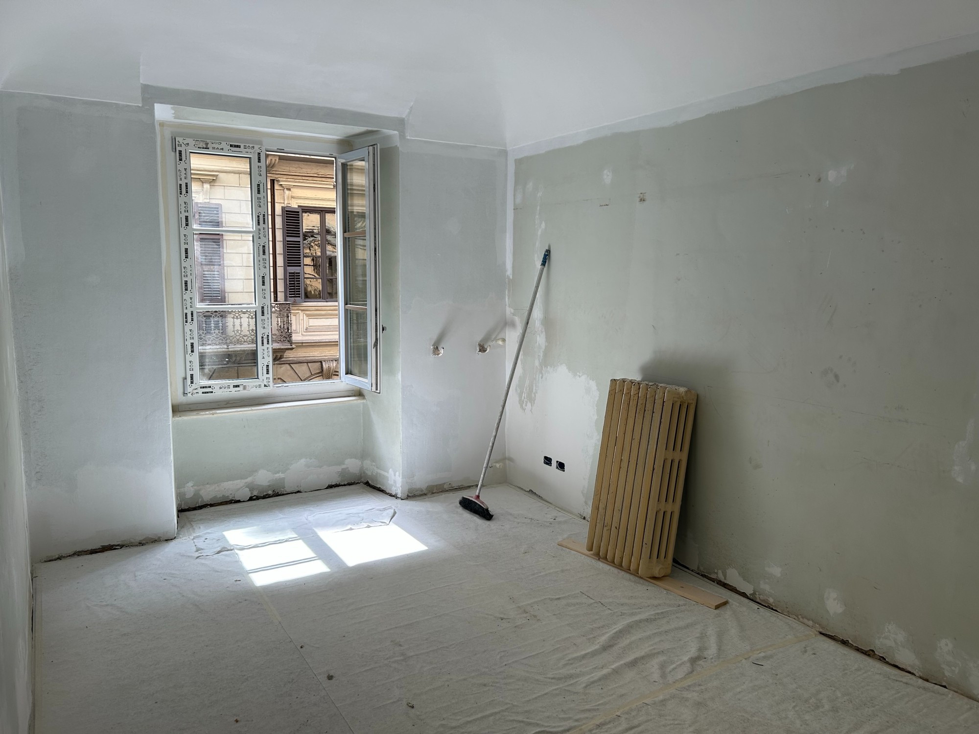 photo of an unfinished apartment room mid-renovation 2023. There is a broom and unhooked radiator against the wall, and the window is open.