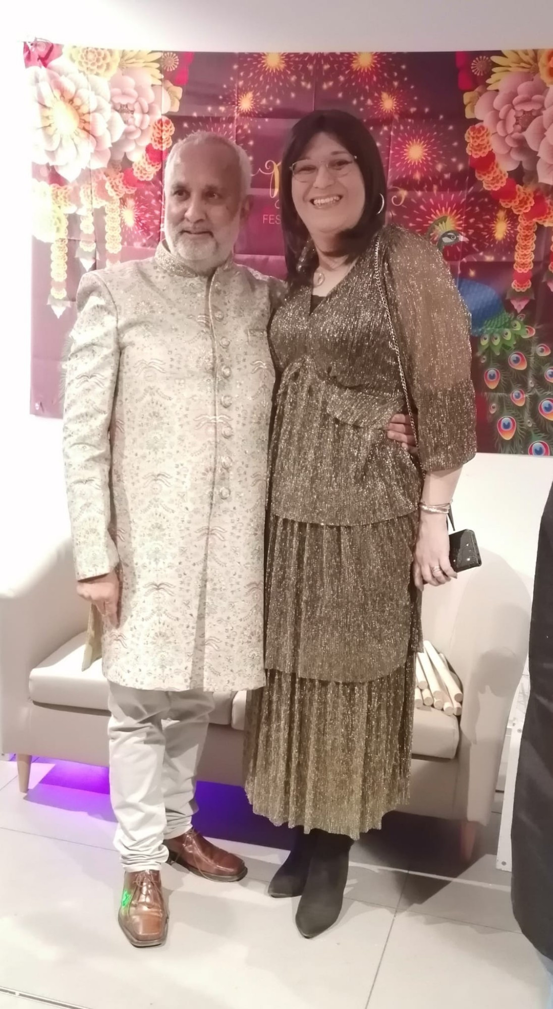 Indian man in white & gold traditional dress standing with a white trans woman in a gold dress in front of a sign that says Happy Diwali