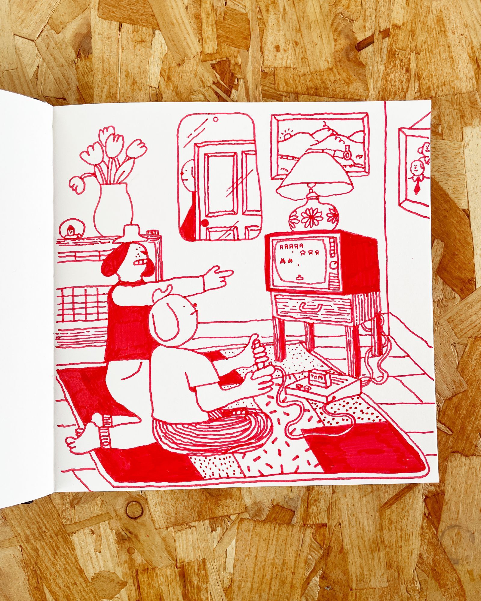 Photo of a sketchbook page with a drawing in red felt tip pen on it. The drawing is of two young animal people sitting on a rug on the living room floor. One is playing Space Invaders on a games console, the other is pointing excitedly at the TV screen.