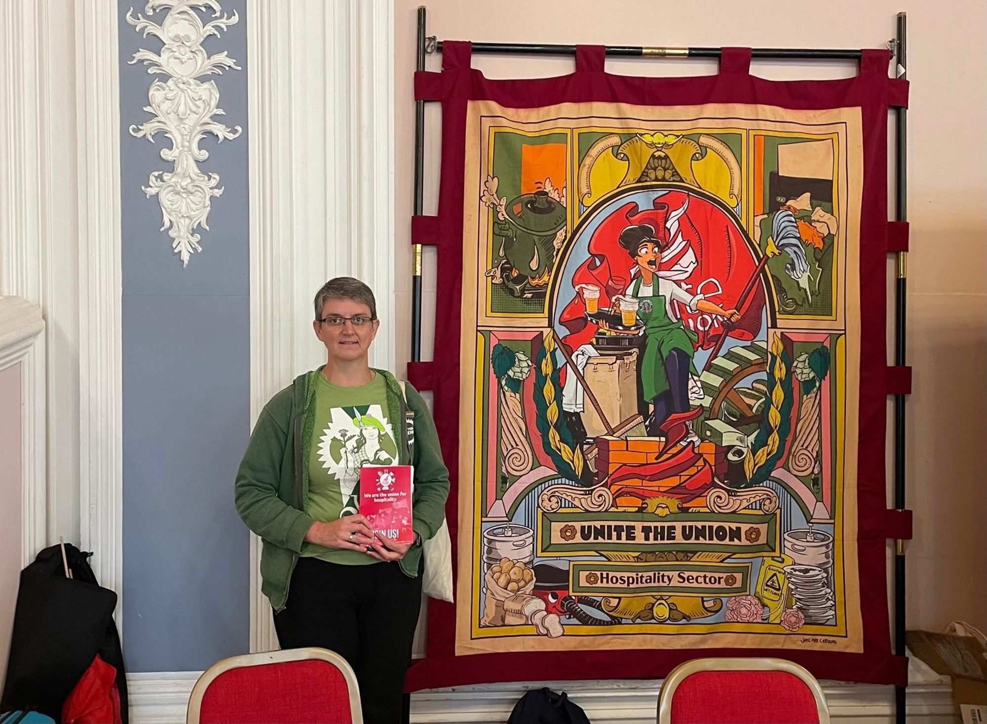 Maggie holding a red unite leaflet next to the hospitality banner which has colourful images of workers and tools on it.