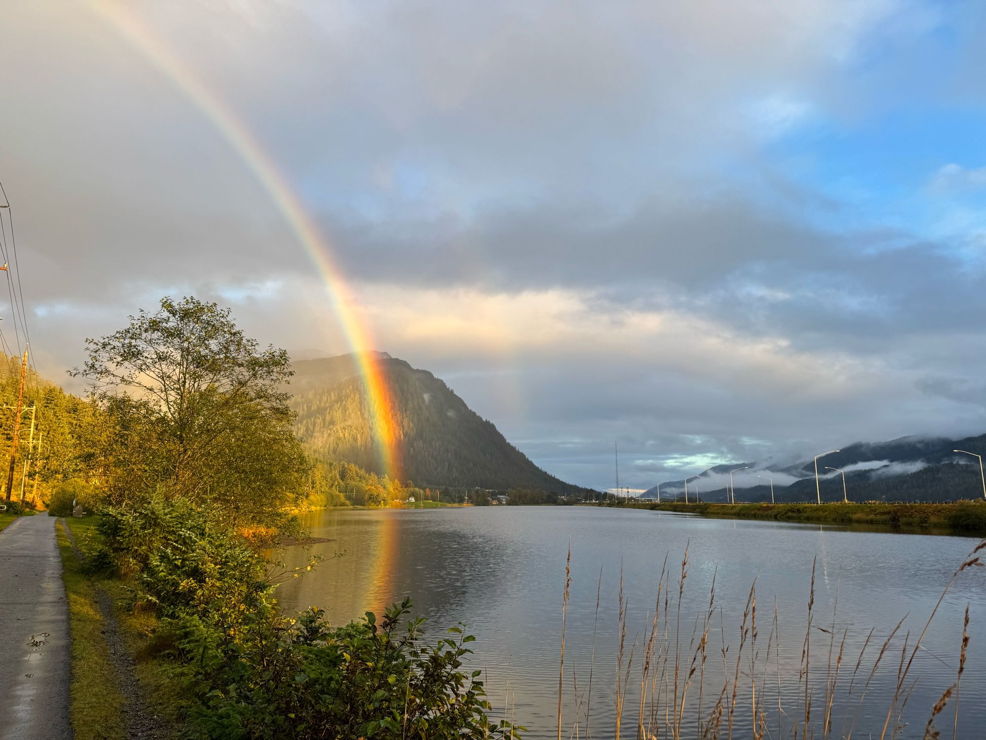 A brilliant rainbow is reflected in a placid lake a mid cloudy skies with patches of blue.