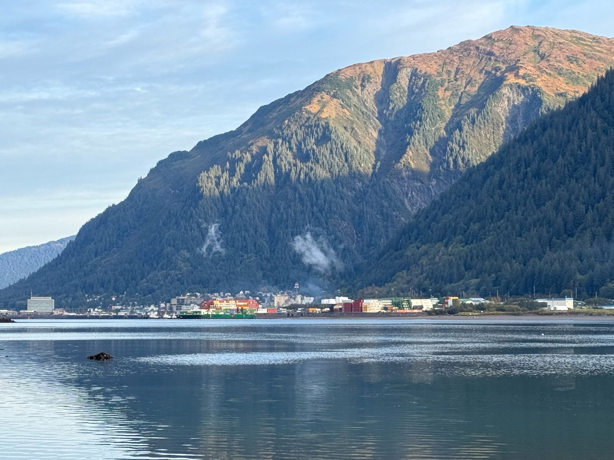 The rising sun illuminates the top of Mount Juneau, with the town below in shadow and a cargo barge preparing to leave