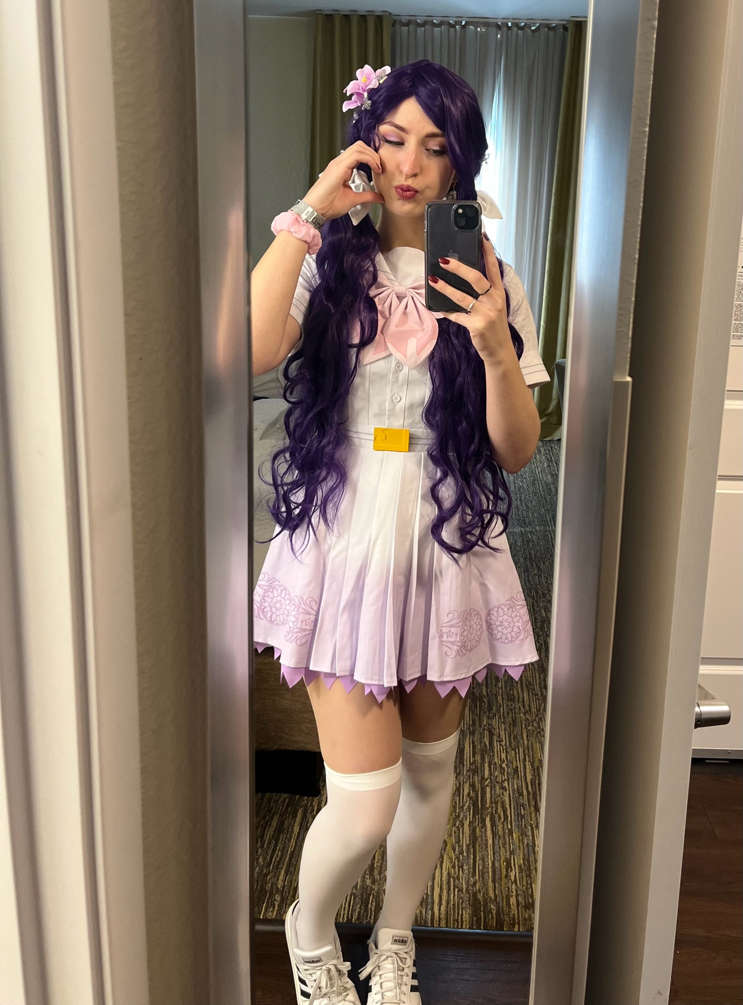 A mirror selfie of Melon dressed as Nozomi Tojo from Love Live: Muse. They’re wearing the Song for you you you! set (a white uniform with a pink neck bow, pale purple hem and little gold belt medallion). There are light purple flowers in their wig.