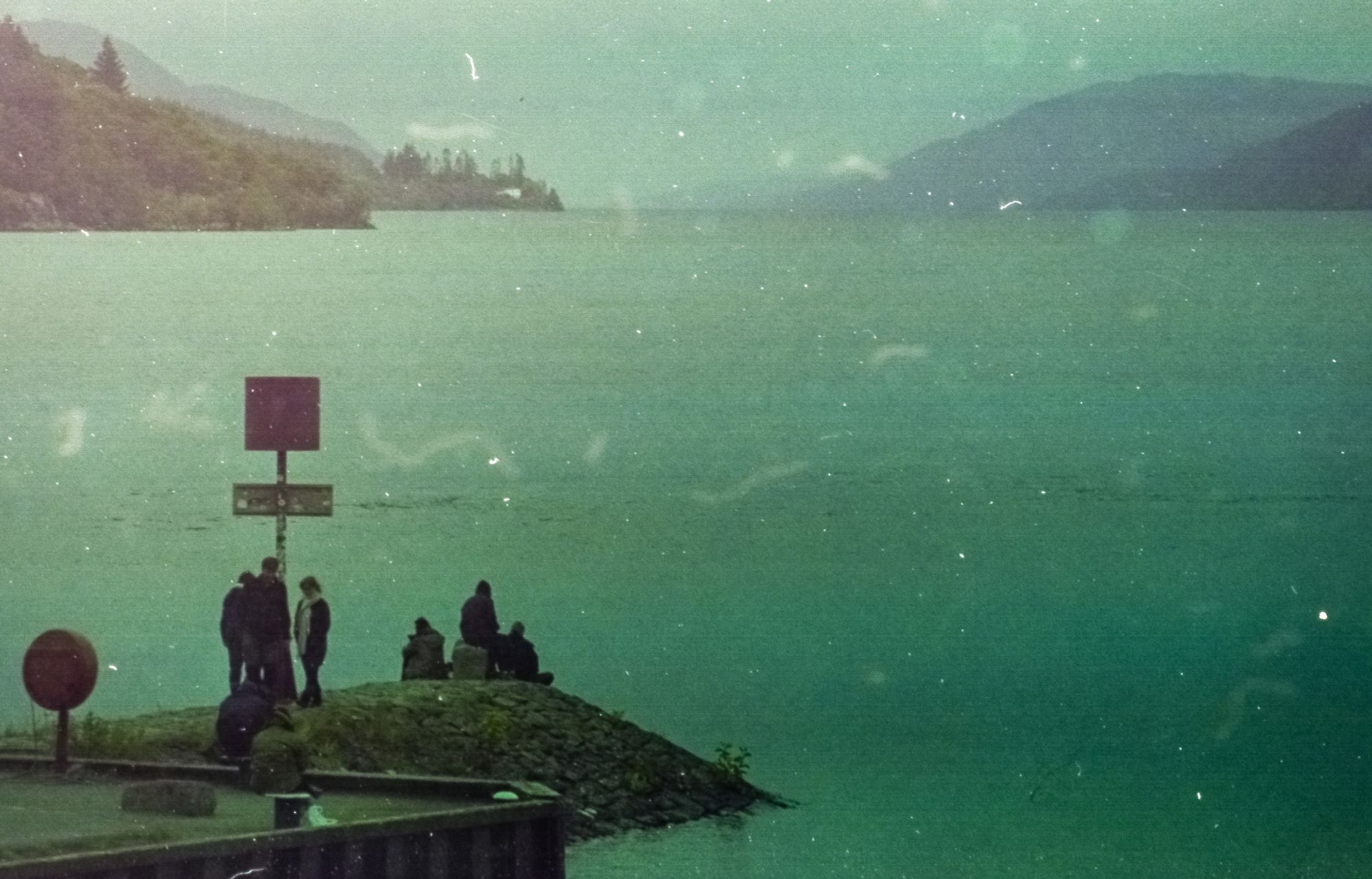 People stand and sit at a jetty looking out onto Loch Ness. This is an analog film Photograph with a green tint.