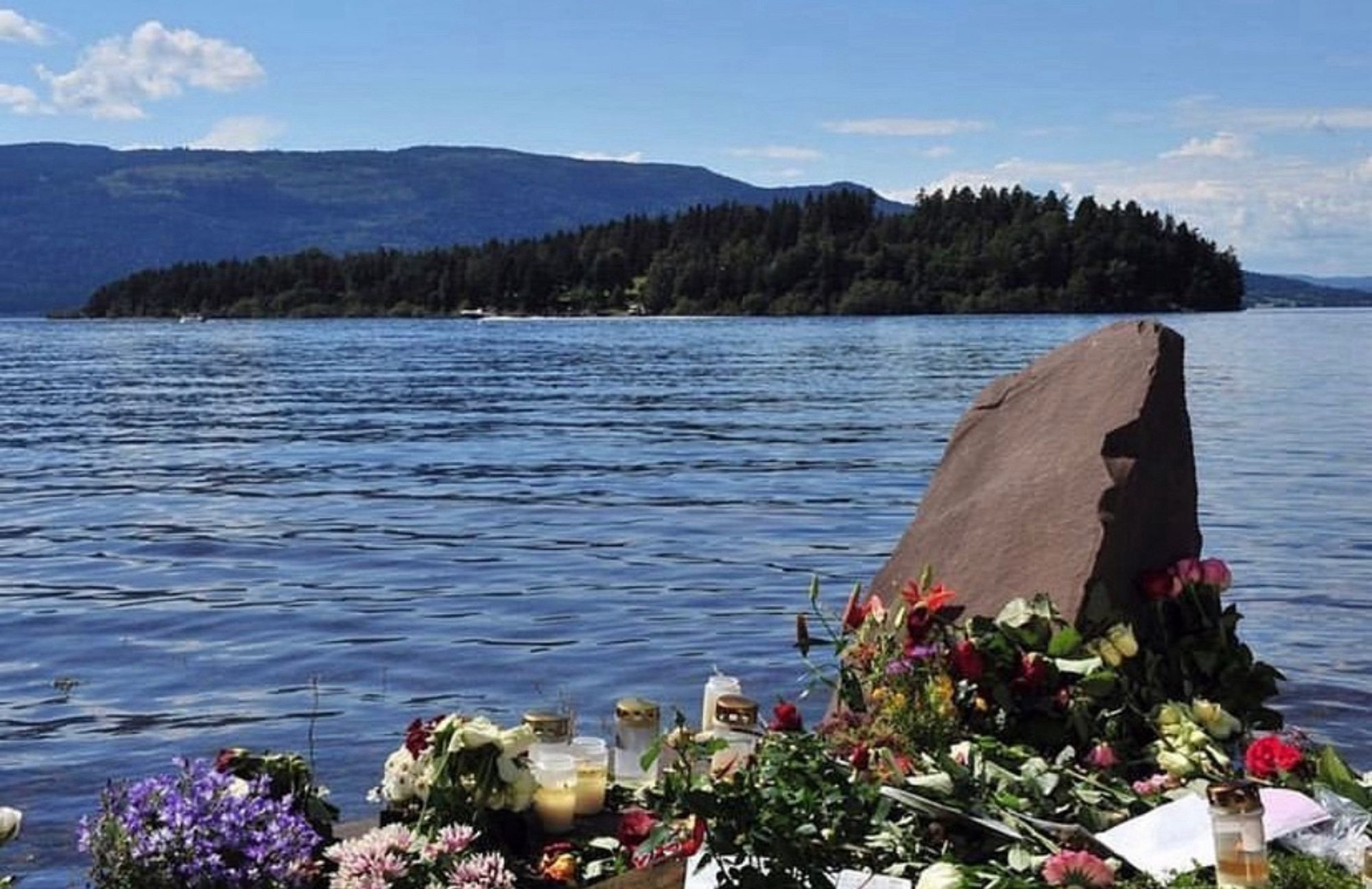 Ein großer Stein am Wasser. Vor dem Stein liegen Blumen. Im Hintergrund erblickt man die Insel Utøya