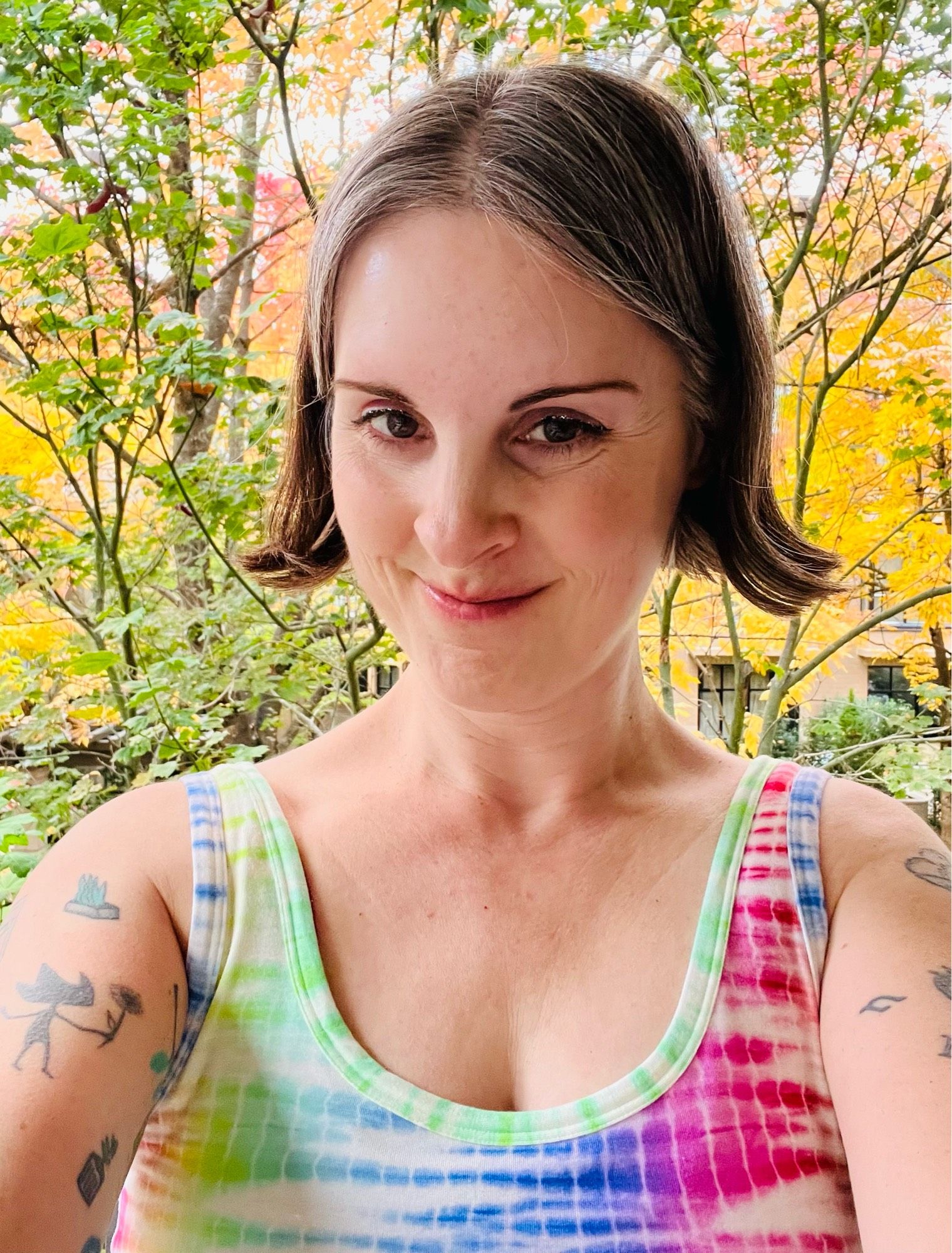 Selfie of Jen wearing a tie dye tank top standing on a balcony with trees and fall leaves behind her.