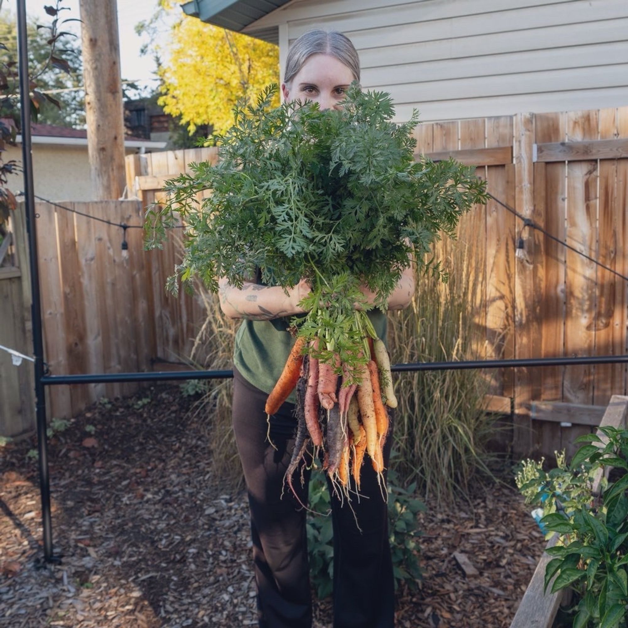 Photo of Jen in the backyard wearing a green T-shirt and black yoga pants holding a bunch of freshly picked garden carrots in front of her like a large bouquet.