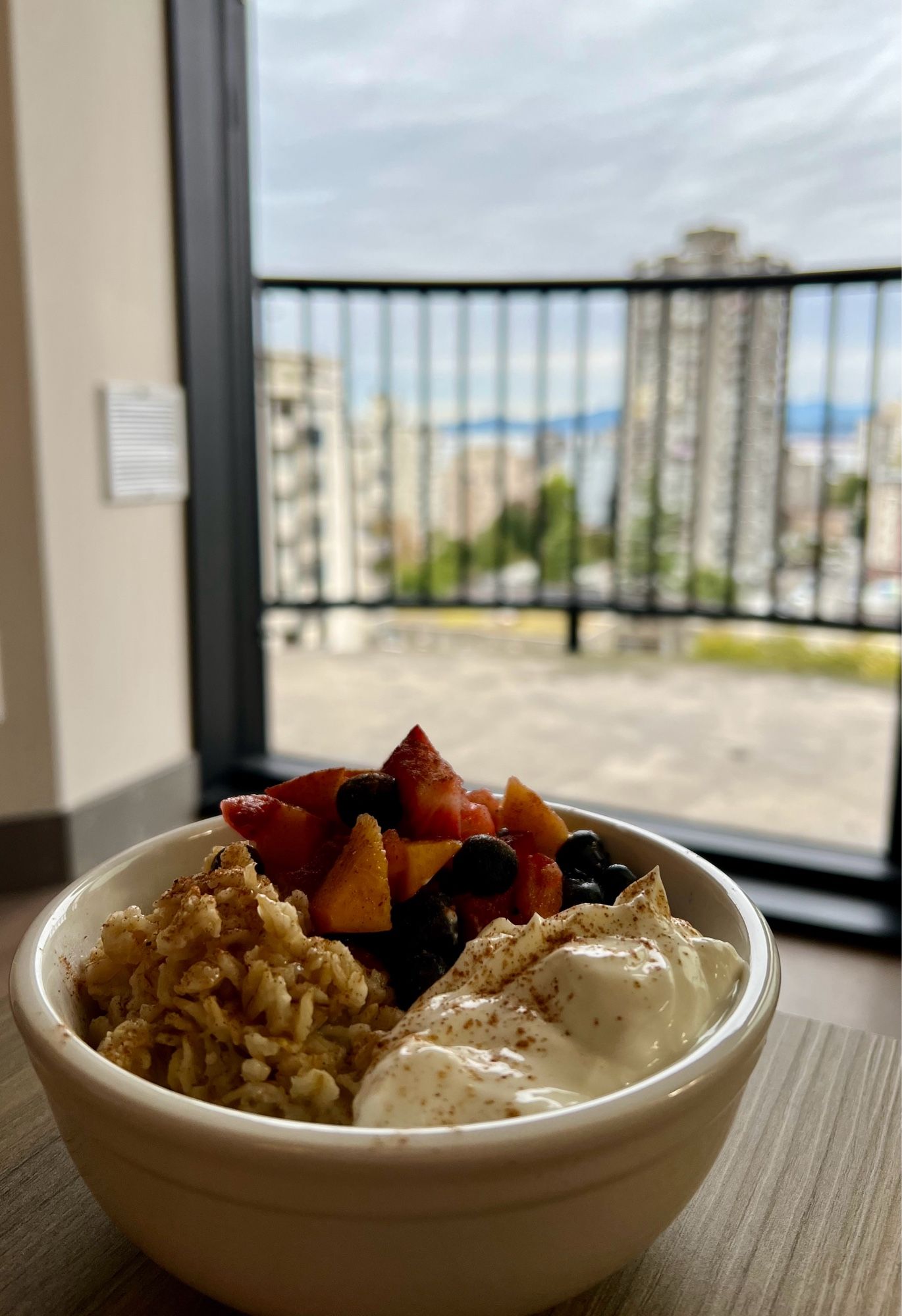 Photo of a bowl of oatmeal, fruit and yogurt sitting on a table next to a hotel balcony with a view overlooking Vancouvers west end.