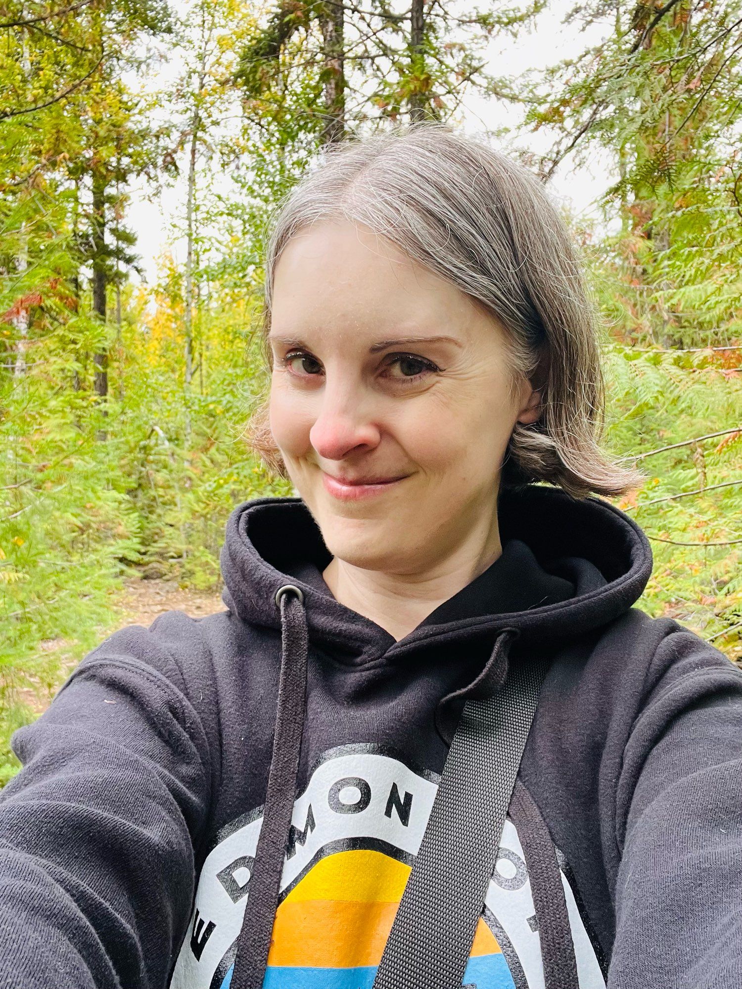 Selfie of Jen wearing a blank hoodie with Edmonton design standing with a beautiful green forest behind her.